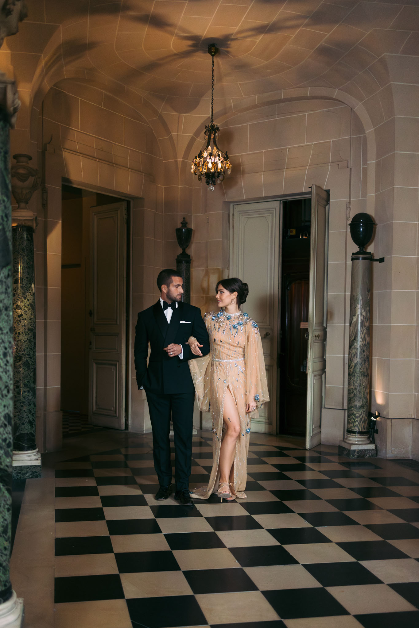 A couple walks arm-in-arm through an indoor grand hallway of what appears to be a French château, set against a black-and-white checkered marble floor beneath a vaulted stone ceiling with a small ornate chandelier. The groom wears a black tuxedo with a bow tie and white pocket square, while the woman wears a champagne-gold gown with flutter sleeves, a front slit, and blue crystal embellishments across the shoulders and bodice, paired with clear heeled sandals and an upswept hairstyle. The corridor features green marble columns, classical arched doorways, and decorative urns on pedestals, creating a formal classical interior setting. The shot is a full-length portrait taken at mid-distance, capturing both figures in motion with warm ambient lighting.