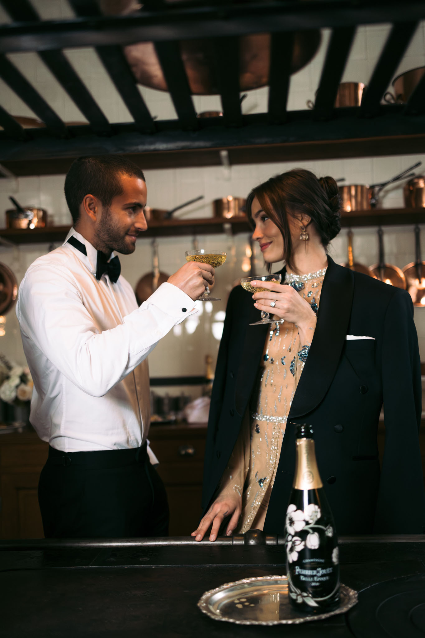 A couple toasts with coupe champagne glasses in what appears to be a vintage kitchen or scullery setting, with copper pots and pans displayed on open shelving along the walls and hanging from a dark ceiling rack above. The groom wears a white dress shirt with a black bow tie and black trousers, while the bride wears a nude embellished gown with teal and silver beading and sequin detailing, draped in a black double-breasted tuxedo jacket; she wears drop earrings and a diamond ring is visible on her left hand. A bottle of Perrier-Jouët Belle Époque champagne sits on a silver tray on the dark countertop in the foreground. The shot is a medium portrait-style image with warm ambient lighting, styled in a formal, black-tie aesthetic.