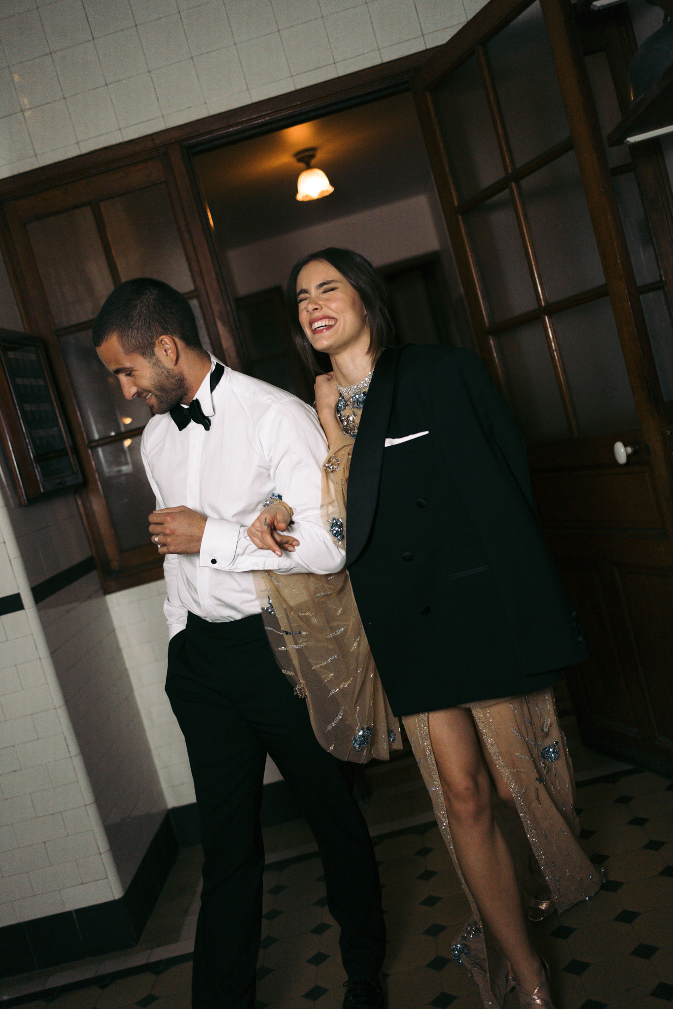 Couple laughing in tiled corridor, woman in champagne beaded dress with groom's tuxedo jacket over shoulders