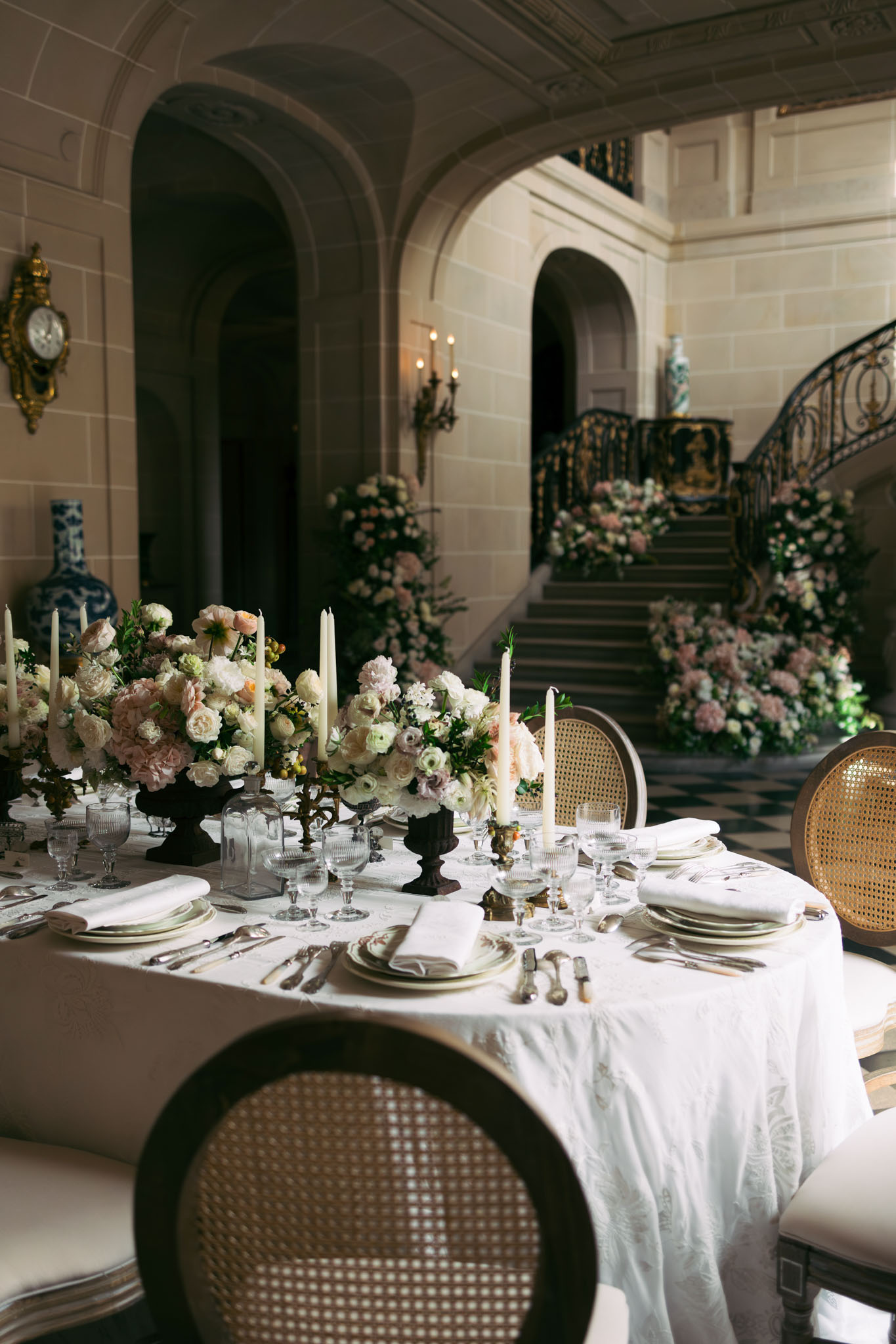 Styled round reception table with blush floral centerpieces and cascading staircase flowers in chateau hall