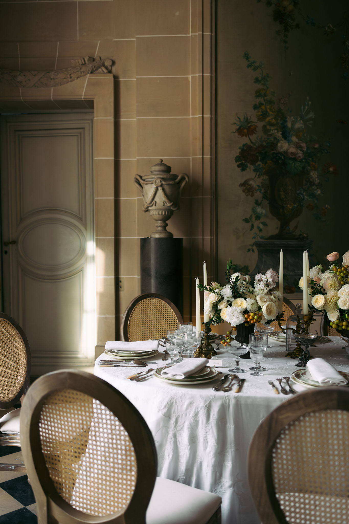 A reception table styling detail shot taken indoors in what appears to be a French château room featuring carved stone walls, a large painted floral mural, a classical stone urn on a pedestal, and a black-and-white diamond-patterned floor. The round table is covered in a white linen cloth and set with stacked plates in muted sage and cream tones, white linen napkins, vintage-style silverware with horn handles, and cut crystal glassware. The centerpiece consists of cream garden roses, white peonies, blush ranunculus, soft lilac flowers, and clusters of small orange berries, arranged in a low floral composition flanked by ivory taper candles in dark antique candelabras. Seating consists of Louis XVI-style cane-back chairs in natural wood. The overall decor palette is ivory, cream, and warm gold with a classic French interior aesthetic. Potential venue feature image.