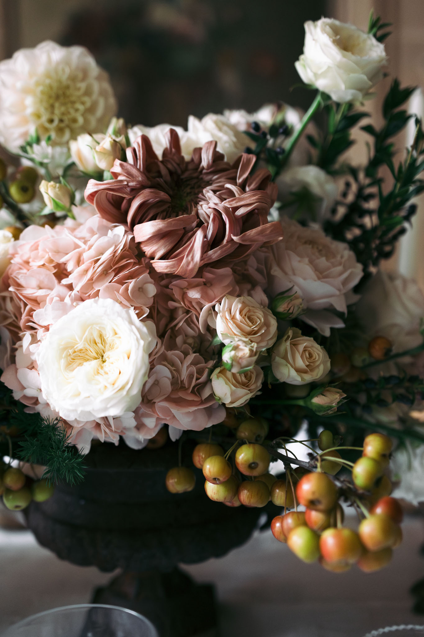 Close-up detail shot of a wedding floral centerpiece arranged in a dark, pedestal-style urn vase. The arrangement features a large mauve-brown dahlia as the focal point, surrounded by blush hydrangeas, cream garden roses, champagne spray roses, white lisianthus, and an ivory pompom dahlia visible in the background. Textural accents include dark berry branches, feathery green foliage, and cascading clusters of crab apples in yellow-green and orange-red tones draped over the edge of the vase. The overall palette is warm and autumnal — dusty rose, terracotta, cream, and champagne — consistent with a classic or romantic autumn wedding aesthetic.