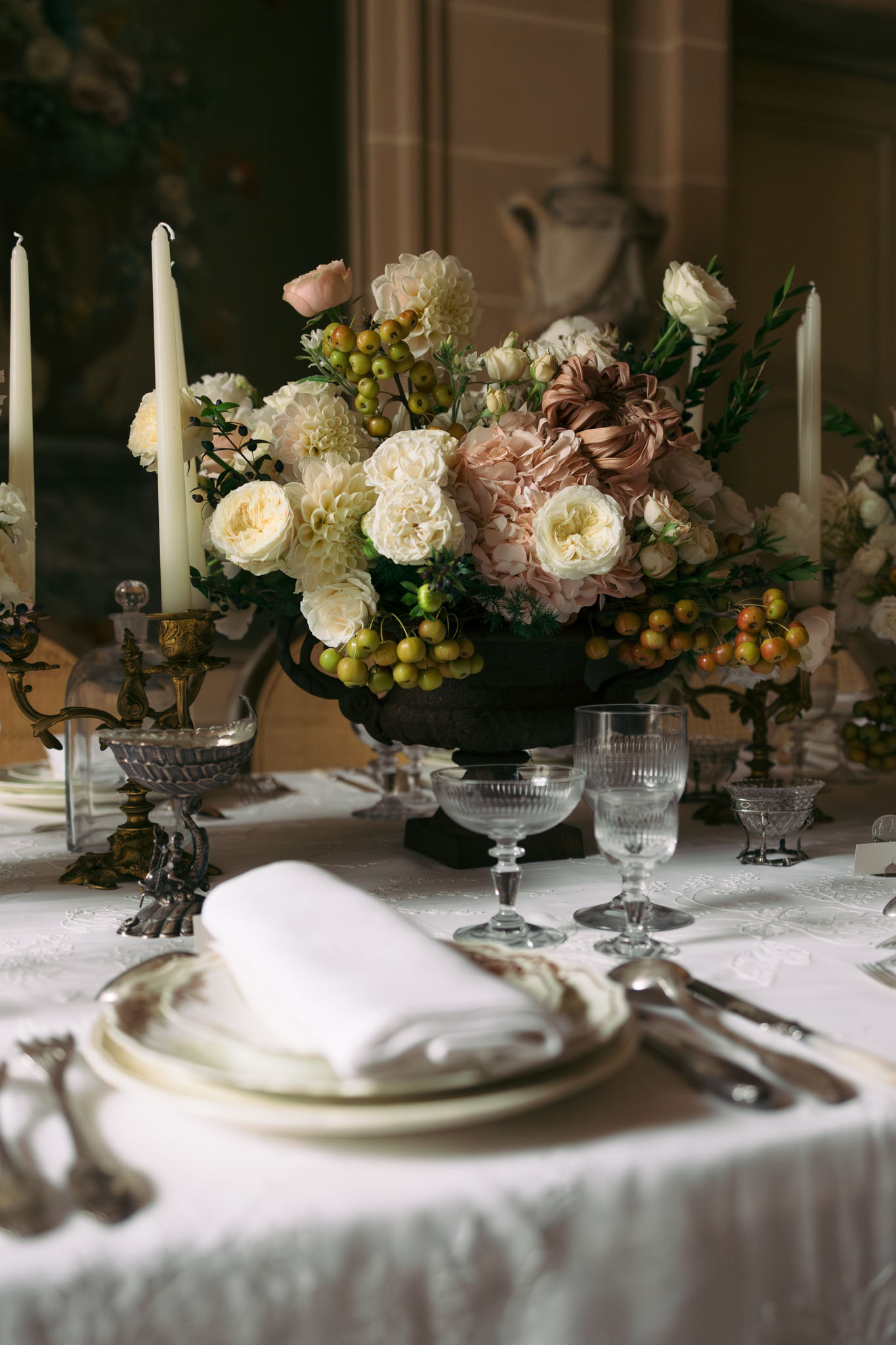 A close-up detail shot of a wedding reception table setting inside what appears to be a château or historic interior, with a painted mural visible in the background. The centerpiece is a dark iron urn filled with a lush arrangement of cream garden roses, blush and mocha-toned dahlias, blush hydrangeas, small white spray roses, green crab apples, and dark foliage. Tall ivory taper candles in ornate antique brass and silver candelabras flank the centerpiece, and smaller matching floral arrangements are visible further down the table. The place settings feature stacked ivory porcelain plates with a folded white linen napkin, silver flatware, and faceted crystal glassware including goblets and coupe-style glasses. The table is dressed in a white damask linen tablecloth, and the overall decor palette is cream, blush, mocha, and antique gold, consistent with a classic or old-world French interior style.