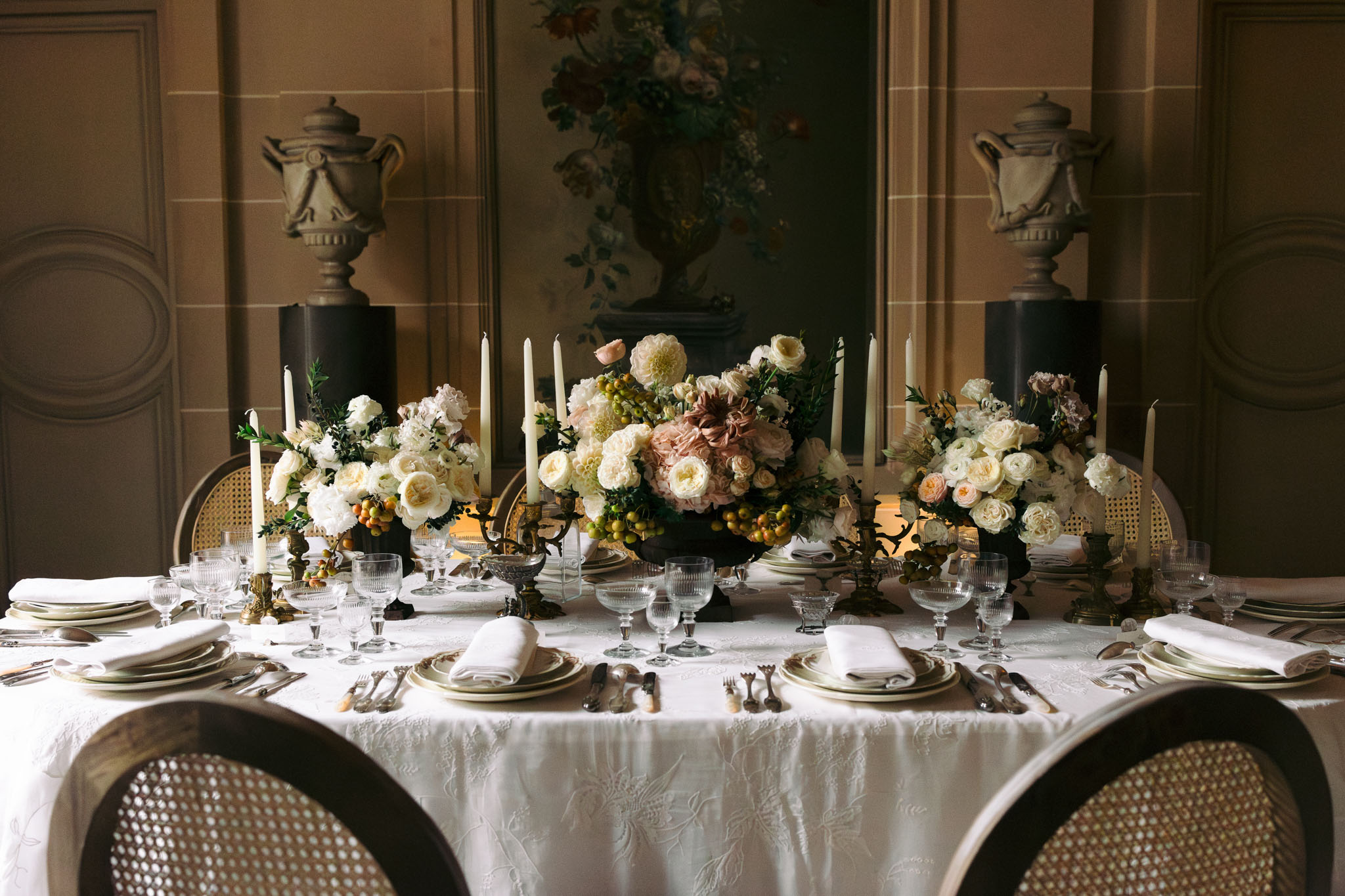 A close-up table styling shot taken indoors in a formal reception room, showing a fully dressed wedding dining table with no guests present. The table is covered in a white damask-patterned linen and set with stacked sage-green and cream plates, white folded napkins, antique-style silver cutlery, and ribbed crystal glassware on brass candleholders. Three low floral centerpieces in dark bowls feature ivory garden roses, cream dahlias, blush peonies, dusty mauve dahlias, small yellow-green berries, and dark foliage, arranged in a lush, abundant style. Tall ivory taper candles are clustered among the arrangements on ornate brass candelabras. The background features warm taupe wood-paneled walls with decorative moulding, two large stone urns on dark pedestals, and a large oil painting depicting a floral arrangement. Cane-back chairs with dark wooden frames are visible in the foreground. The overall palette is ivory, blush, dusty mauve, brass, and warm taupe, with a classic French interior style.