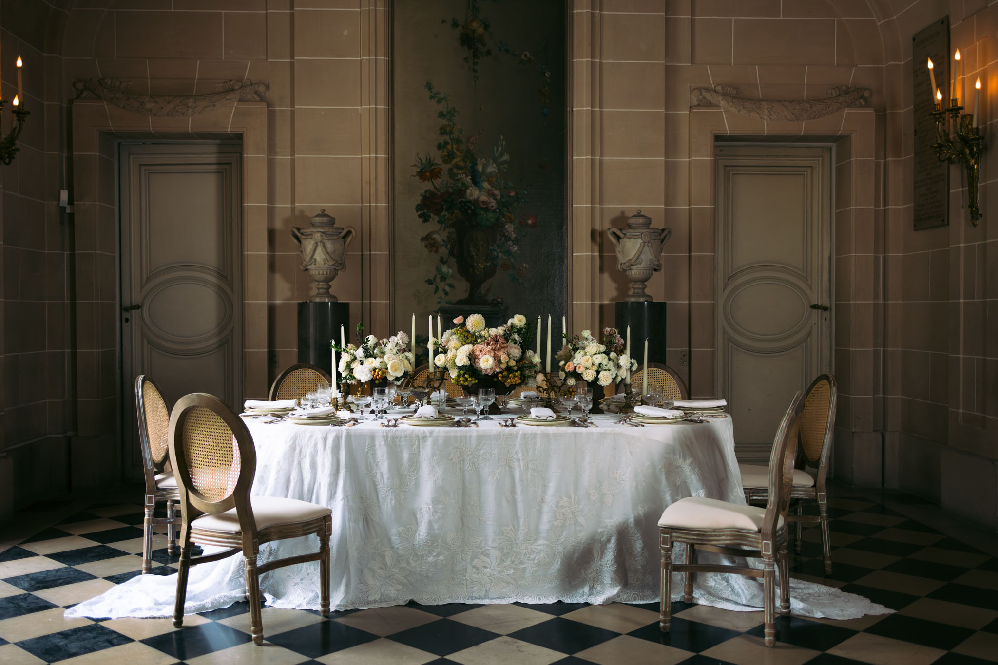 A reception table styling shot inside a grand French château room, with no guests present. The oval table is covered in a white jacquard-patterned linen and set for approximately eight guests, with cream linen napkins, gold-rimmed charger plates, crystal glassware, and ornate silverware. The centerpiece consists of lush floral arrangements featuring cream and blush garden roses, dusty mauve dahlias, ivory hydrangeas, and small clusters of green berries, interspersed with tall ivory taper candles in brass holders. Louis XVI-style cane-back chairs with cream upholstered seats surround the table. The room features stone-block paneled walls, classical painted stone urns on pedestals flanking a large oil painting of a floral bouquet on the back wall, bronze wall sconces with lit candles, and a black-and-white diamond-pattern marble floor. The overall decor palette is warm ivory, blush, taupe, and aged gold with a classic French interior aesthetic. Wide, symmetrically composed shot. Potential venue feature image.