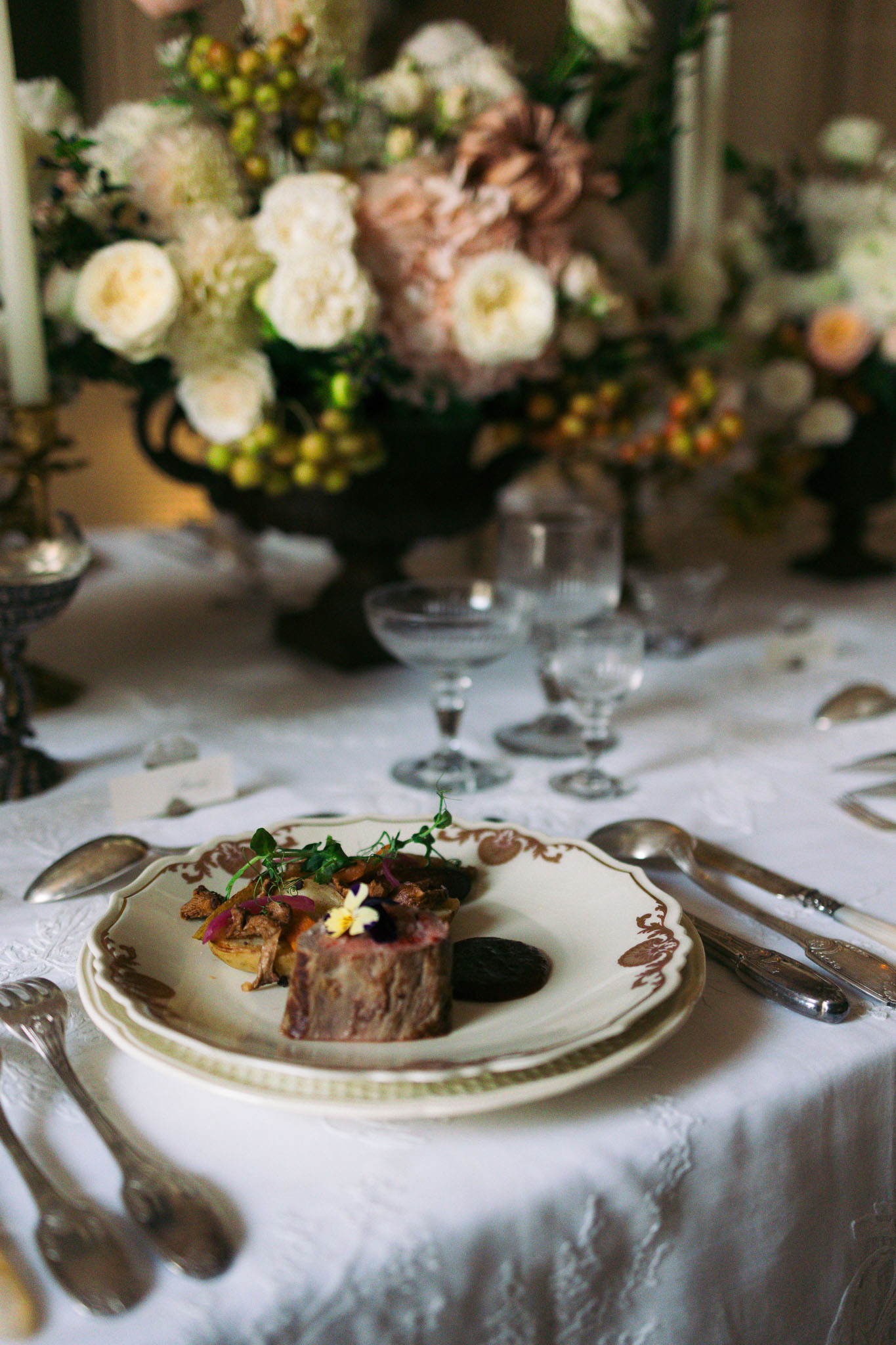Wedding reception table setting with plated main course on antique porcelain, cream roses and mauve dahlia centerpiece