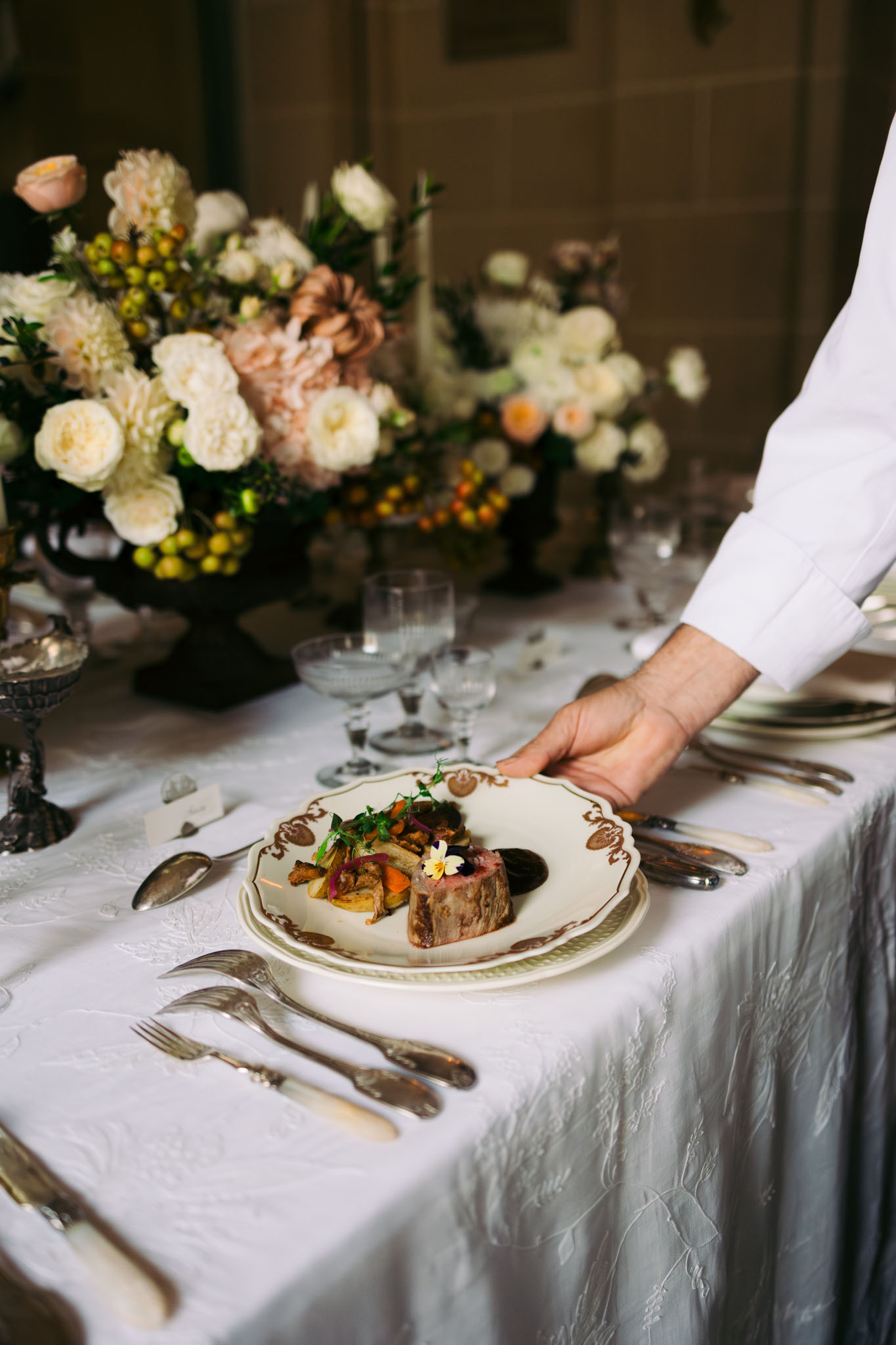 Server placing plated main course on gold-trimmed china at formal reception table with rose and dahlia centerpieces