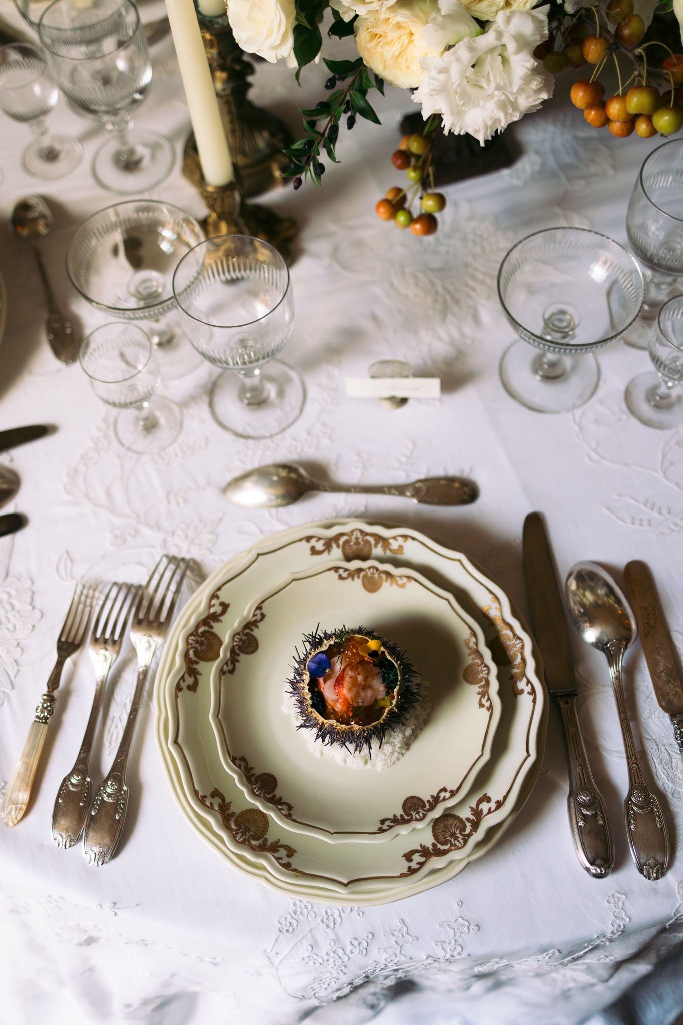 Wedding reception place setting with gold-bordered plates, sea urchin appetizer, crystal glassware, and peony centerpiece