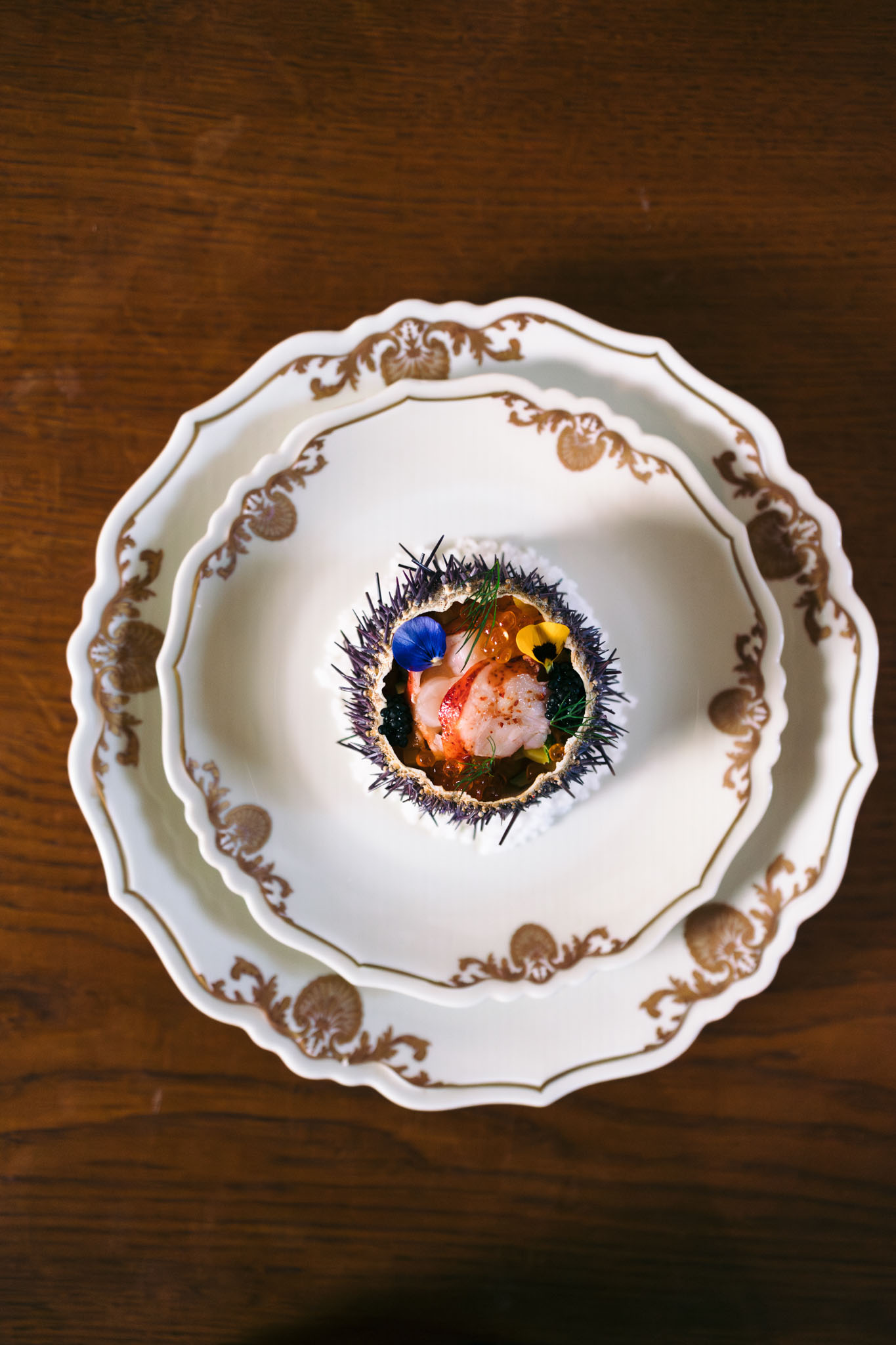 A close-up overhead shot of a wedding reception catering detail, showing a fine dining starter course served inside an open sea urchin shell. The dish is plated on two stacked white porcelain plates with ornate gold baroque-style border detailing and scalloped edges, set on a dark wood surface. The sea urchin shell contains what appears to be lobster or prawn, black caviar, salmon roe, fresh dill, and is garnished with a small blue edible flower and a yellow edible flower petal, with a light dusting of red spice. The composition is a portrait-orientation flat lay, with sharp focus on the dish highlighting the high-end catering style chosen for the reception menu.