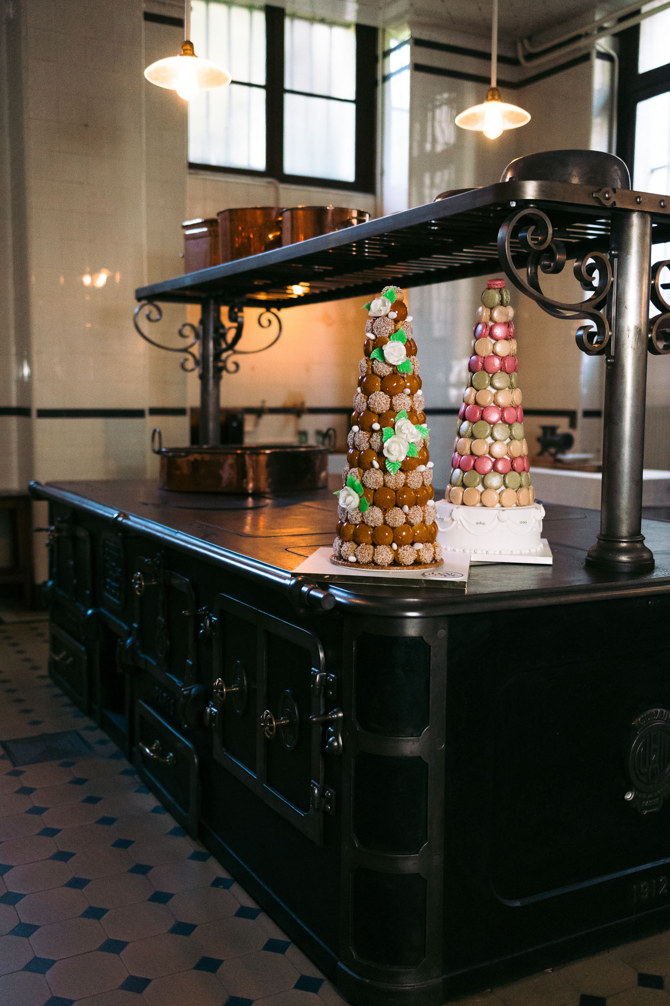 A detail shot of two French wedding cakes displayed on top of a large black cast-iron professional kitchen range with ornate wrought-iron detailing and a geometric blue-and-white tiled floor. The taller cake on the left is a traditional croquembouche — a tower of caramel-glazed choux puffs decorated with white sugar flowers and green fondant leaves. Beside it sits a conical macaron tower arranged with macarons in shades of dusty rose, sage green, beige, and pistachio, placed on a white cake stand. The kitchen setting features copper pots in the background, pendant lights with cream globe shades, and white tiled walls with black trim detailing, giving a classic French culinary institution atmosphere.