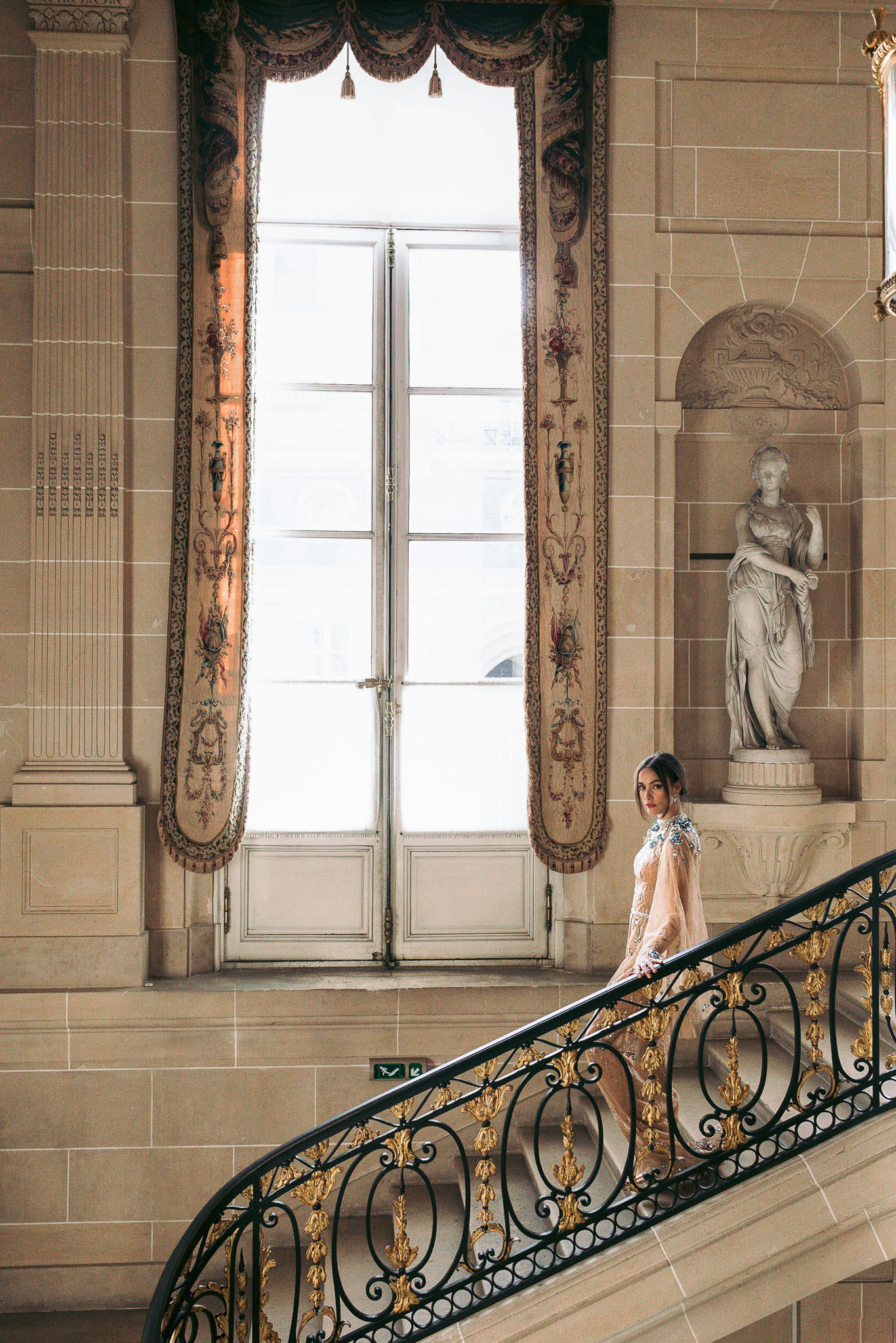 Bride in embellished blush cape gown on grand staircase with gilded iron balustrade and classical architecture