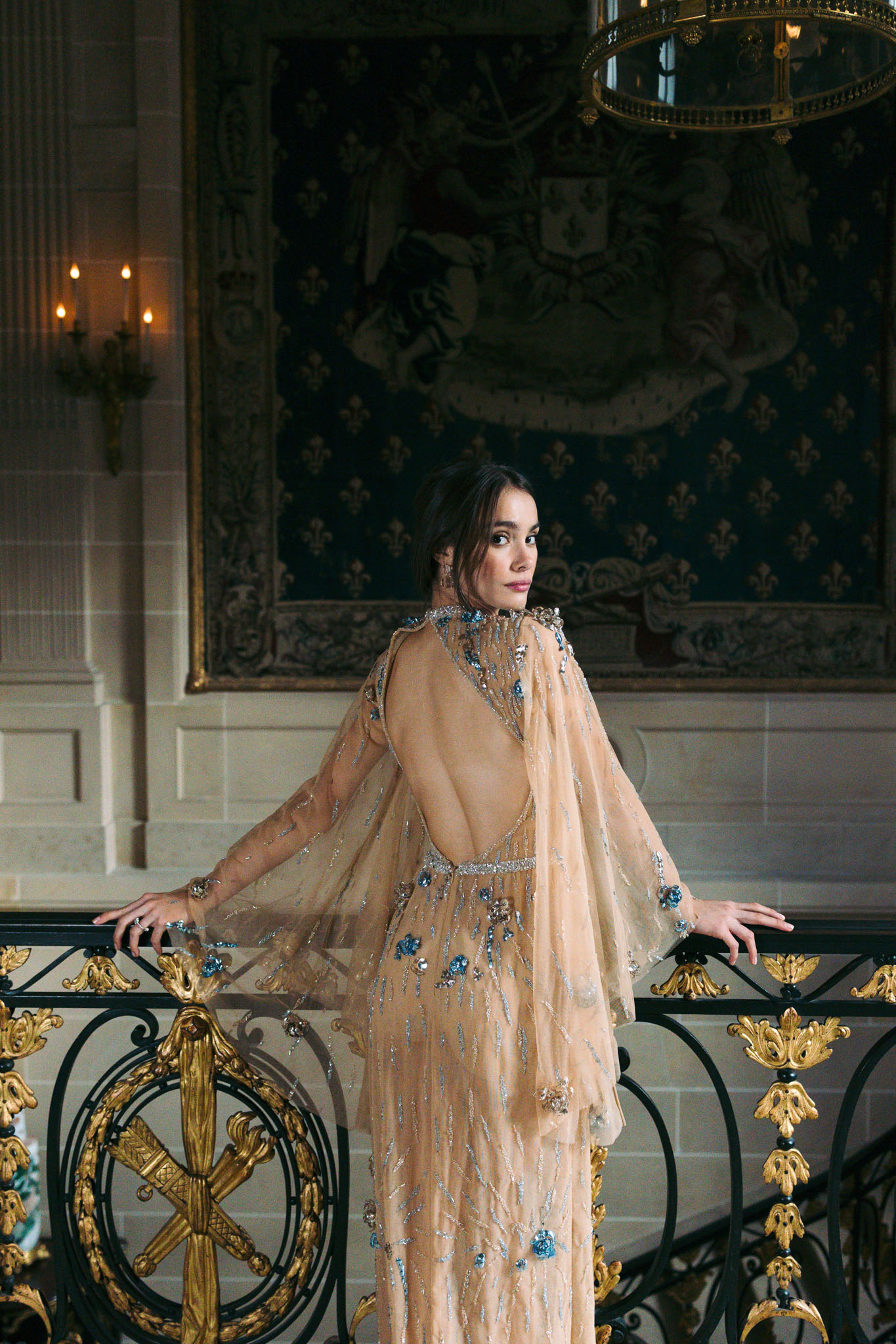 A portrait of a bride posing on an ornate interior staircase balcony, looking back over her shoulder toward the camera. She wears a champagne-nude gown with a deep open back, cape-style sleeves, and heavy embellishment including silver beading and blue crystal floral appliqués throughout the sheer overlay. The setting is a grand château interior featuring a large decorative tapestry with heraldic motifs in deep teal and gold behind her, a gilded and wrought-iron balustrade with gold leaf detailing in the foreground, and a brass chandelier visible overhead. Wall sconces with lit candles are visible to the left, contributing to warm, low ambient lighting. The styling is formal and ornate, consistent with a classic French château aesthetic. Medium portrait shot, slightly from behind.