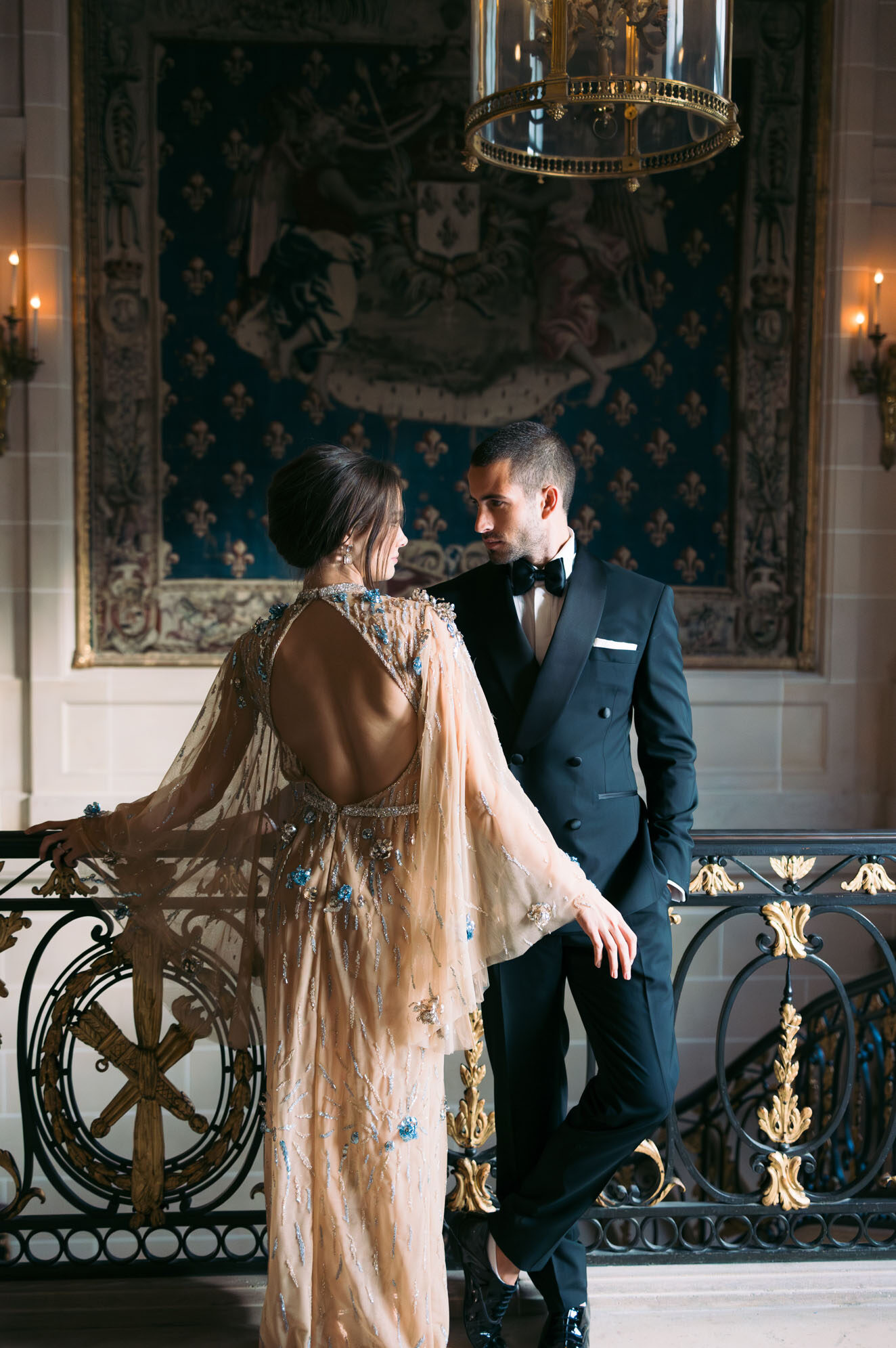 A couple poses on an ornate grand staircase landing inside a château or historic palace-style venue, facing each other in an intimate portrait composition. The bride wears a champagne-gold heavily embellished gown with an open back, sheer cape sleeves, and blue floral beaded details throughout; her hair is swept up. The groom wears a navy double-breasted tuxedo with a black bow tie, white pocket square, and patent leather shoes. Behind them hangs a large royal-blue heraldic tapestry with gold fleur-de-lis motifs, framed in gilt, and the balustrade features black wrought iron with ornate gold-leafed scrollwork. Warm candlelight from wall sconces and a brass-and-glass ceiling lantern illuminate the scene, reinforcing a formal, palatial aesthetic. Potential venue feature image.