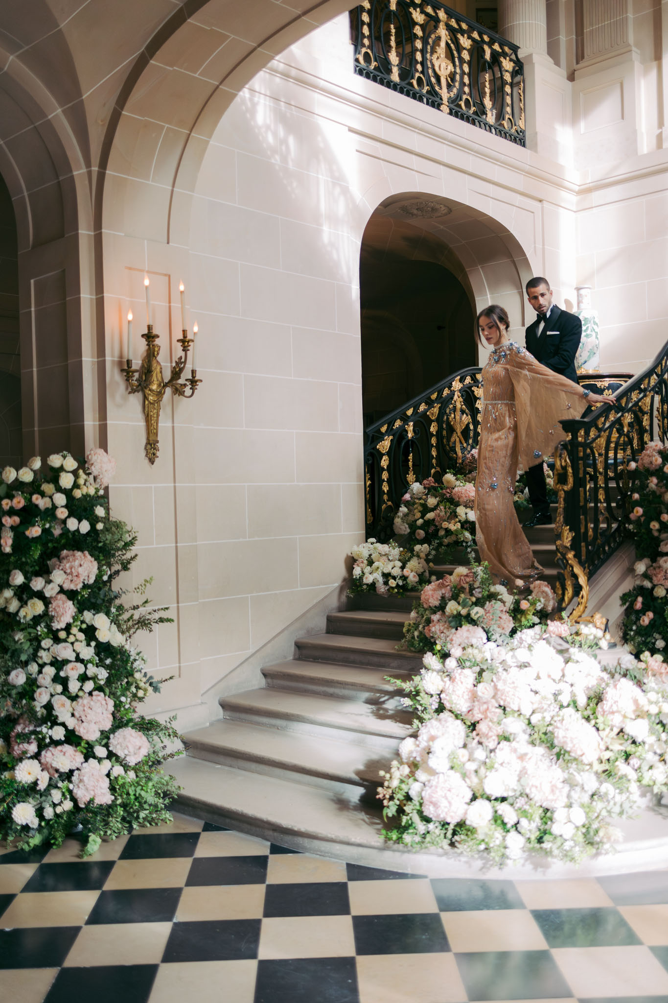 A couple poses on a grand interior staircase of what appears to be a French château or palace-style venue, shot from a wide-angle perspective. The woman wears a champagne-gold embellished gown with a sheer cape overlay featuring blue crystal details, while the man is dressed in a black tuxedo. The staircase is decorated with large floor-level floral installations composed of blush pink hydrangeas, ivory garden roses, blush dahlias, and lush greenery, cascading along both sides of the steps and pooling at the base. The interior features a black-and-white marble checkered floor, arched stone doorways, ornate black-and-gold wrought iron balustrades, a gilded candelabra wall sconce, and a decorative black-and-gold upper balcony railing, all contributing to a classic, formal French interior aesthetic. Potential venue feature image.