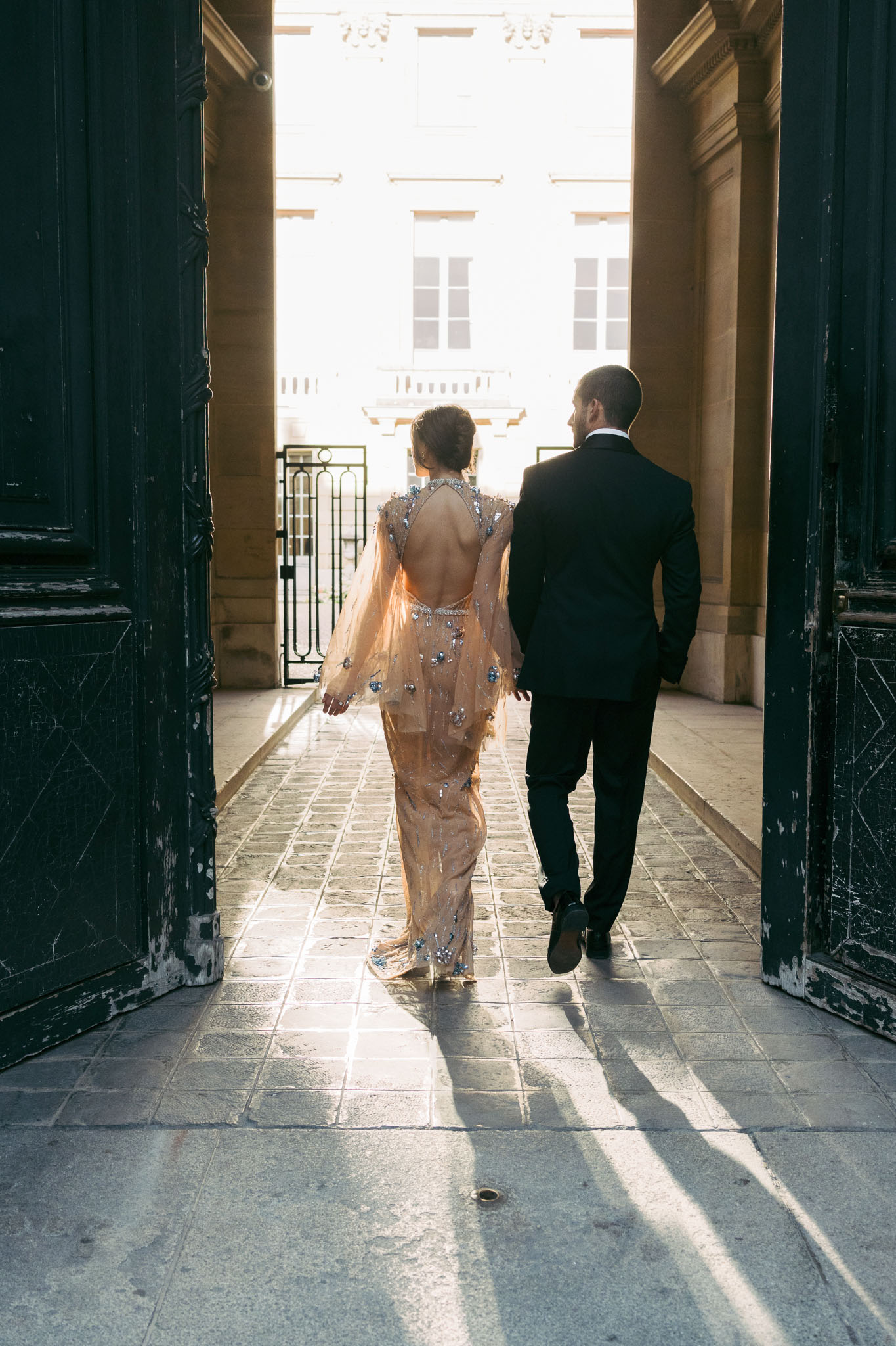 A couple portrait shot from behind as the bride and groom walk together through large dark green double doors into a sunlit Haussmann-style Parisian courtyard. The bride wears a champagne-peach sheer embellished gown with an open back, long sheer sleeves, and scattered crystal and blue gemstone detailing throughout; the groom wears a fitted black tuxedo suit. The composition frames the pair centrally between the doors, with strong backlighting creating long shadows across the stone tile floor and silhouetting the classical French building facade visible beyond the iron gate in the background. The shot is a full-length walking portrait with a modern, fashion-forward styling aesthetic.