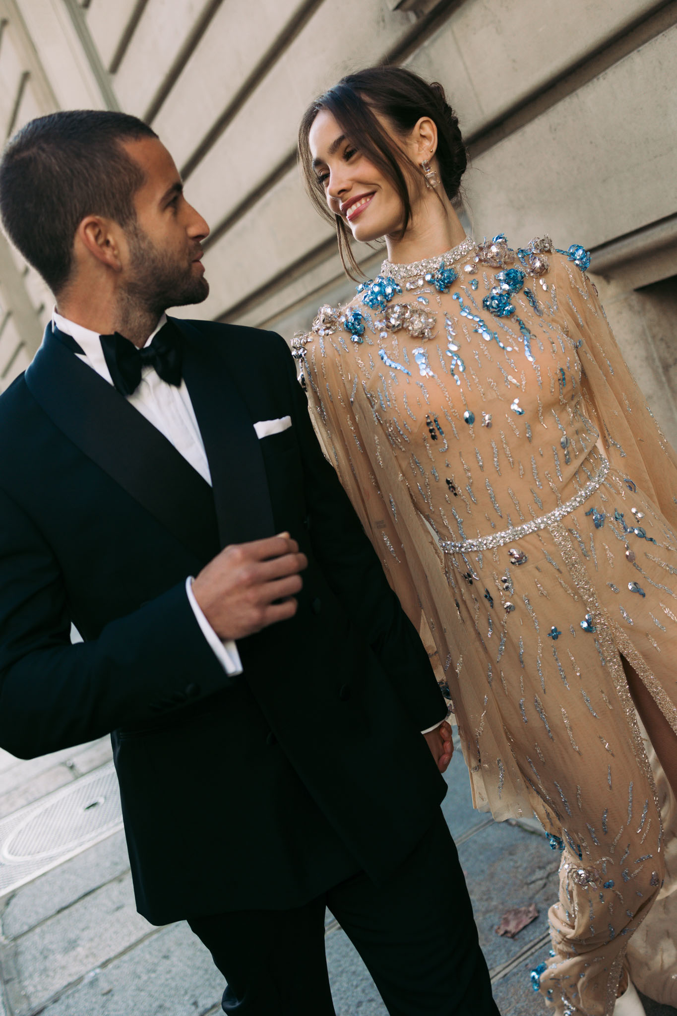 A couple portrait shot outdoors against a large stone architectural facade. The groom wears a black tuxedo with a black bow tie, white dress shirt, and white pocket square. The bride wears a champagne-nude, heavily embellished gown featuring silver sequin detailing, a crystal-studded waist belt, and three-dimensional blue floral appliqués at the shoulders, with long sheer sleeves and a high neckline. Her dark hair is styled in a loose updo. She is smiling and looking at the groom, while he looks toward her. The composition is a close-up couple portrait with a modern, formal styling aesthetic.