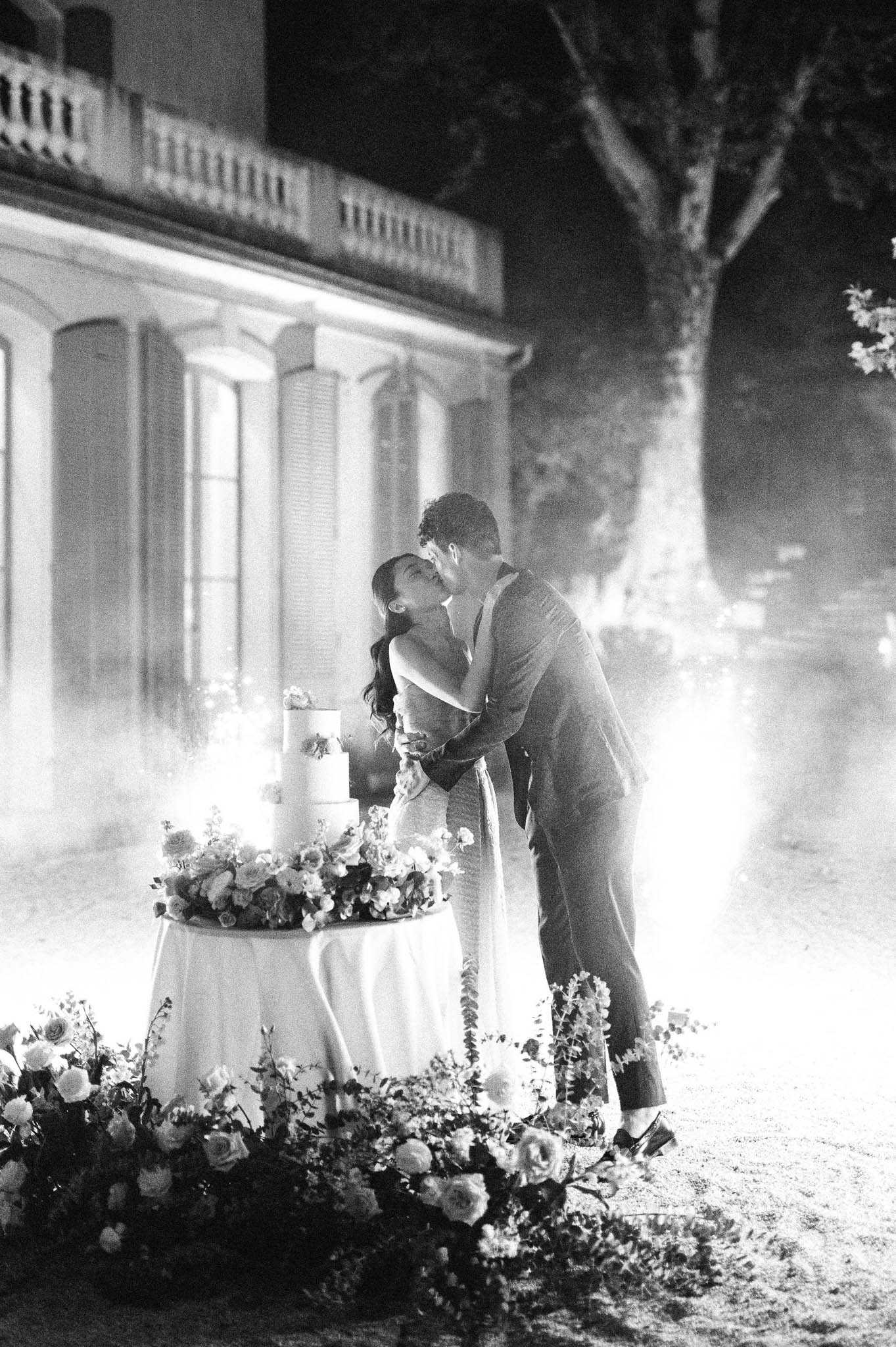 Couple kissing beside tiered cake with sparklers and floral ground display before chateau facade at night in B&W