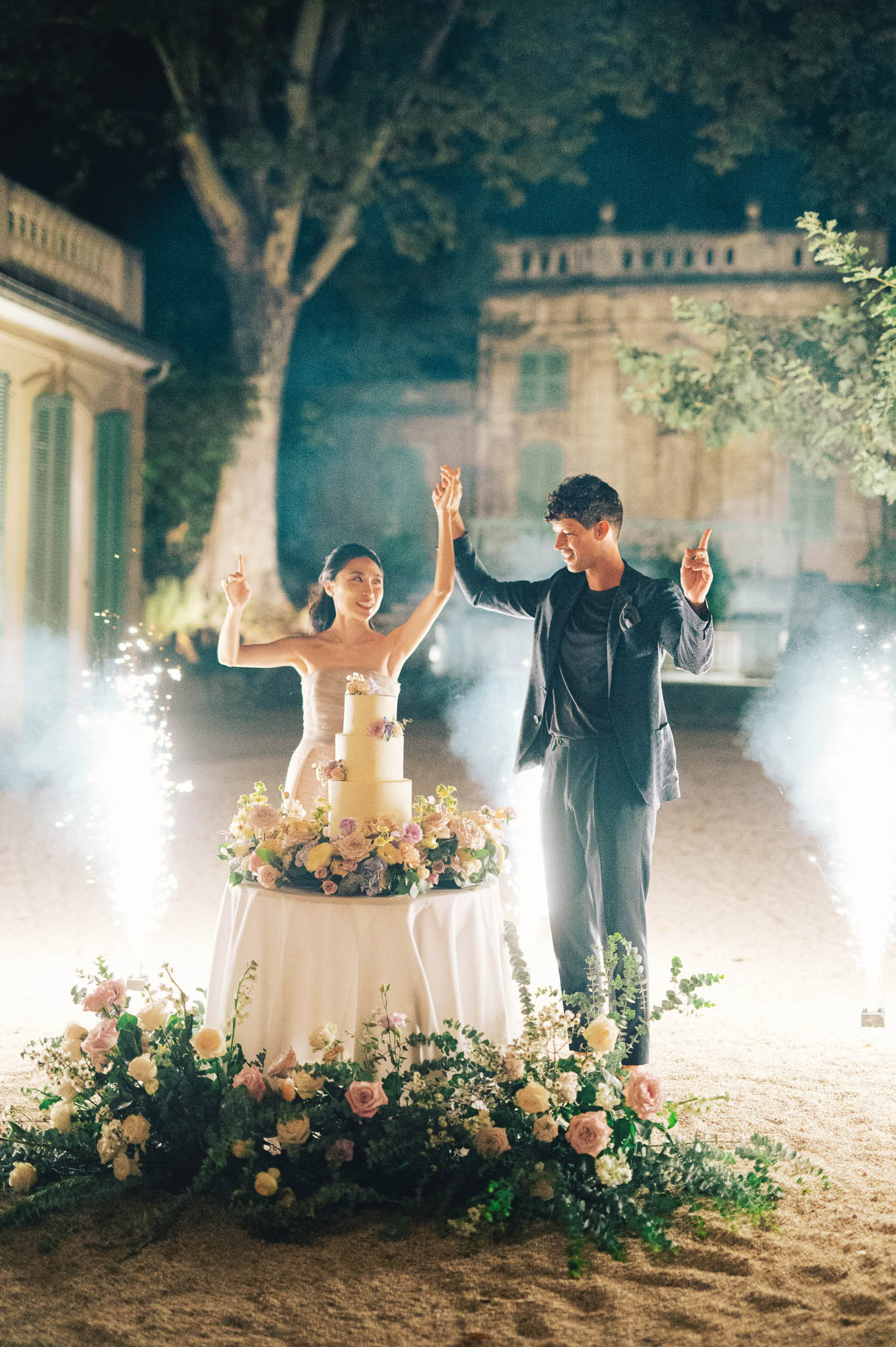 Nighttime cake cutting with sparklers beside four-tier cake decorated with blush and lavender florals