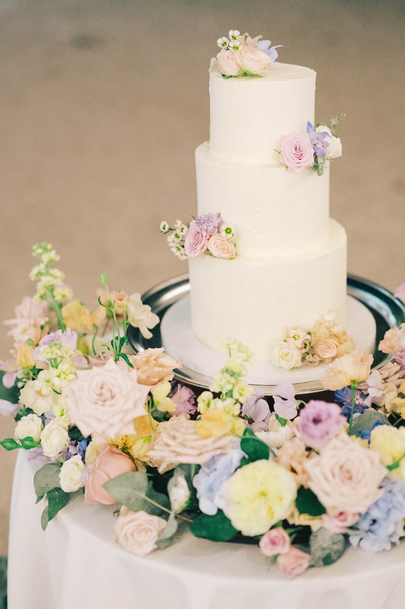 Close-up detail shot of a three-tiered white wedding cake with a smooth buttercream finish, displayed on a silver cake stand atop a white linen-covered table. Each tier is decorated with small clusters of blush roses, soft lavender roses, and white wax flowers. The cake is surrounded by a generous arrangement of loose florals including blush garden roses, peach roses, pale yellow ranunculus, light blue hydrangeas, lavender sweet peas, cream spray roses, and greenery, creating a full garden-style base arrangement. The overall floral palette is pastel and multicolored, consistent with a romantic, garden-inspired aesthetic.