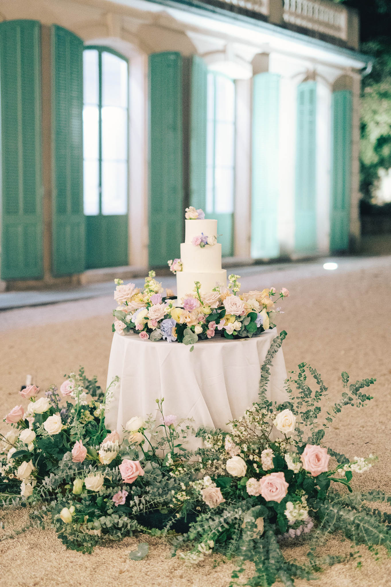 Four-tier ivory cake with lavender blooms surrounded by blush rose ground installation at night courtyard