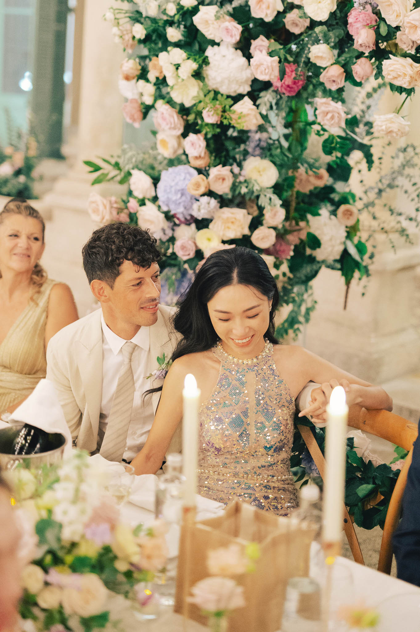 The couple is seated at what appears to be the head table during an indoor wedding reception, likely inside a classical venue with stone architectural elements visible in the background. The groom wears a light beige suit with a striped tie and a small floral buttonhole, while the bride has changed into a halter-neck reception dress featuring gold and iridescent beaded embellishment with a gold beaded neckline; she is smiling warmly with her eyes downcast. Behind them rises a large, lush floral installation composed of blush pink roses, ivory garden roses, white hydrangeas, lavender hydrangeas, deep pink roses, and abundant greenery. The foreground table is styled with tall white taper candles, low floral arrangements in blush and cream tones, and glassware, while a female guest in a gold dress is visible to the left. The shot is a medium portrait-style image capturing the couple from the waist up in a candid, relaxed moment.