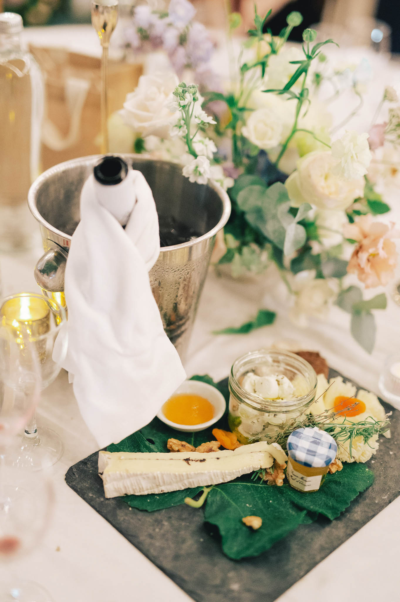 A close-up detail shot of a wedding reception table featuring a French cheese course served on a slate board lined with large green fig leaves. The board holds a wedge of brie-style soft-rind cheese, walnuts, dried apricots, a small jar of honey with a blue gingham lid, a glass jar of herbed fromage blanc or similar fresh cheese, and a small dish of amber-colored jam or honey. A polished silver champagne bucket with a white linen napkin draped over an iced bottle sits immediately behind the board. In the background, a lush floral centerpiece composed of ivory garden roses, blush ranunculus, lavender, and eucalyptus foliage is partially visible alongside champagne flutes and gold votive candles, suggesting a romantic, garden-inspired reception décor palette in soft whites, blush, and green tones.