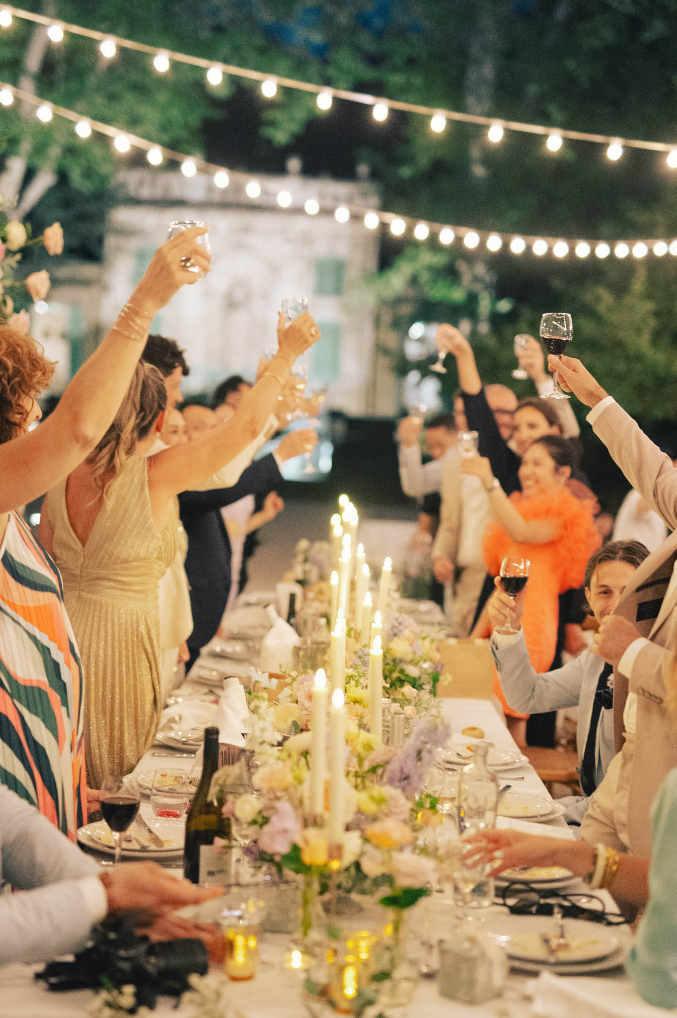 Guests seated and standing along a long banquet table raise their glasses in a toast during an outdoor evening reception. The setting appears to be the grounds of a French château or manor, with an illuminated classical facade visible in the background and bistro string lights strung overhead creating warm ambient lighting. The table is dressed with a lush low centerpiece runner of cream, pale lavender, and soft yellow florals interspersed with clusters of white taper candles in varying heights, alongside wine bottles, glassware, and white dinner plates. Guests are dressed in colorful attire including a bold orange-and-teal striped outfit, a gold sequined dress, and a bright orange feathered or ruffled jacket, giving the reception a festive, eclectic feel. The shot is taken from a low angle along the length of the table, creating a perspective portrait that captures approximately 15–20 guests mid-toast.