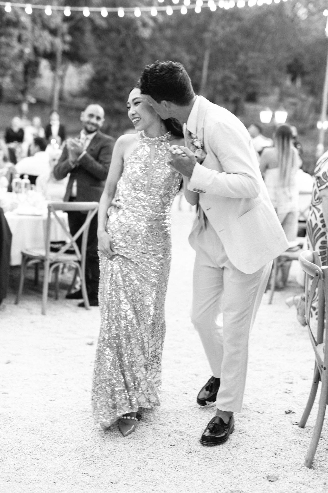 This black-and-white image captures a couple making an entrance at an outdoor evening reception, walking between seated guests on a gravel surface. The bride wears a floor-length sequined halter-neck gown with a pearl necklace and pointed heels with pearl embellishment; the groom is dressed in a light-colored suit with dark loafers and a boutonniere, and is leaning in to kiss her on the cheek as she laughs. String bistro lights are strung overhead, and cross-back wooden chairs with white-clothed tables are visible in the background, where guests are seen applauding. The image is a mid-shot with soft contrast, capturing a candid, joyful moment during the reception grand entrance.