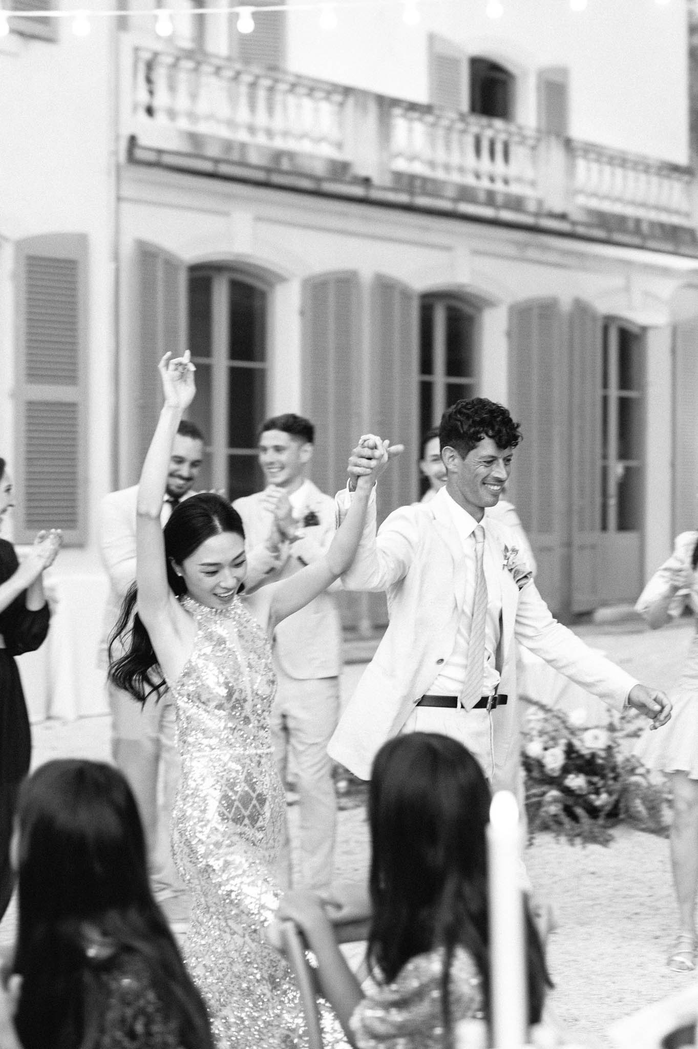 This black-and-white image captures a lively outdoor reception dance on the courtyard of a French château or manor house, identifiable by its shuttered arched windows and wrought-iron balcony in the background. The bride, wearing a heavily embellished sequined or beaded fitted dress, has her arm raised in celebration while holding hands with the groom, who is dressed in a light-coloured suit with a tie and a boutonnière. A string of globe lights is visible overhead, and several guests and bridal party members in light-coloured suits can be seen dancing and clapping behind the couple. The image is shot in high contrast black and white with bright highlights and soft mid-tones, rendered in a medium portrait-style shot with shallow depth of field.
