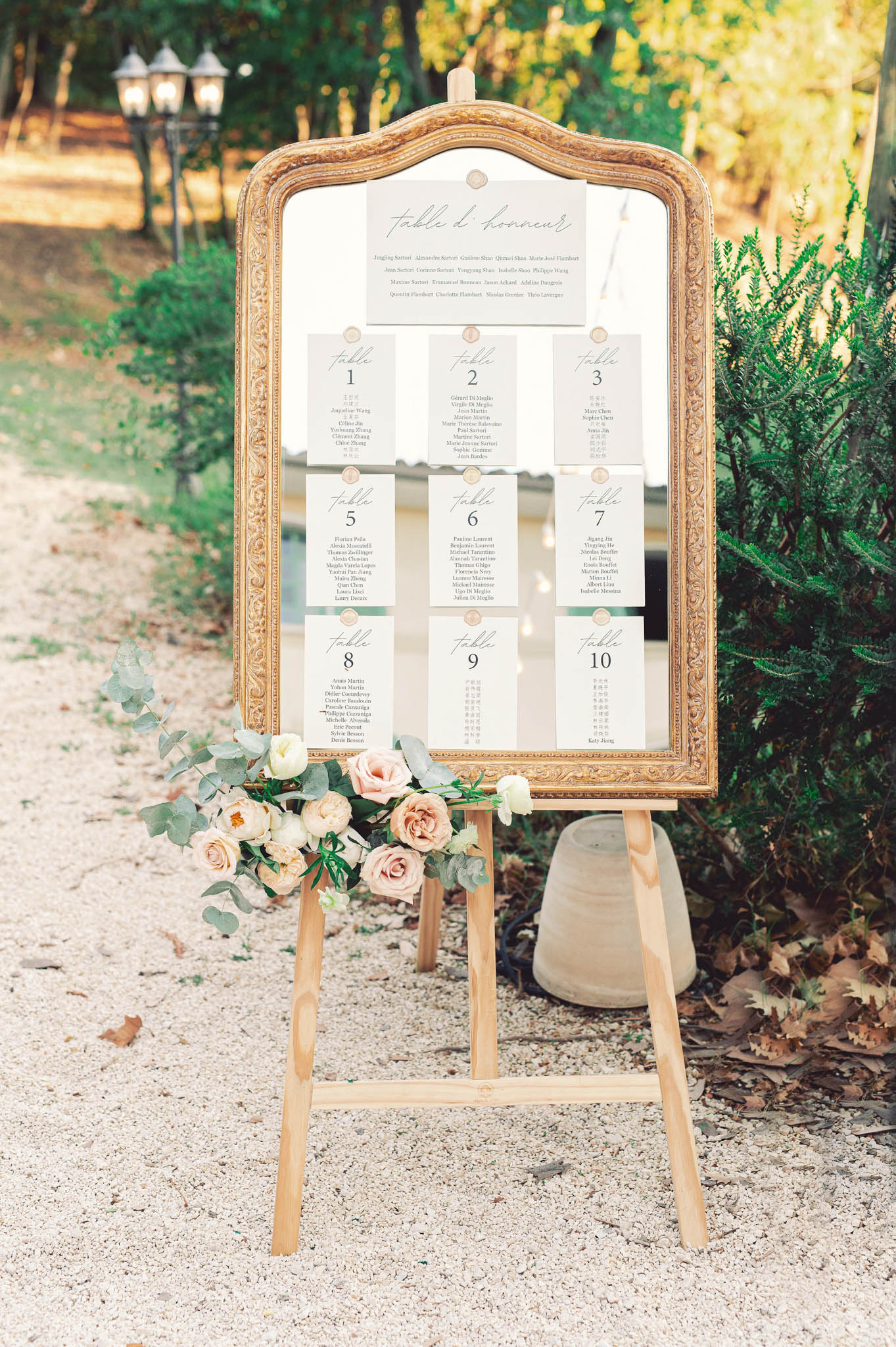 A close-up detail shot of an outdoor wedding seating chart display on a gravel path. A gold ornate mirror frame is mounted on a natural wood easel, holding white card seating plan cards arranged in a grid layout — a 'table d'honneur' header card at the top followed by cards for tables 1 through 10, each listing guest names in a clean script and serif font with a sage green accent stripe. The base of the easel is decorated with a cluster of blush and champagne roses, ivory roses, and eucalyptus sprigs. The overall decor palette is white, gold, blush, and sage green, consistent with a classic French-style wedding aesthetic.