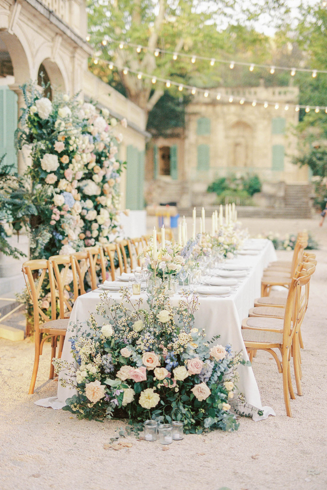Courtyard table with cascading blush rose and delphinium runner ivory tapers Edison lights and floral column at stone buil...