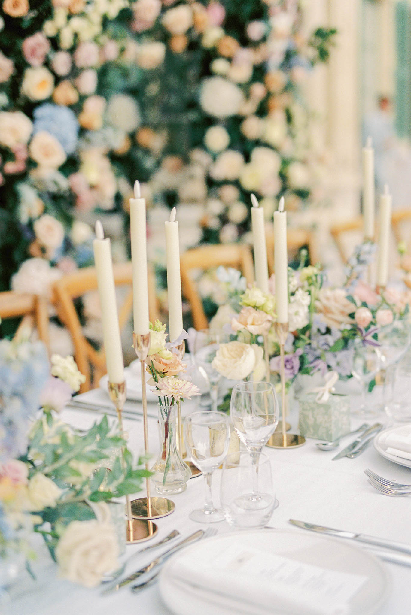 A close-up detail shot of a wedding reception tablescape, shot at a slight angle to show the length of the table. The white linen table is set with white plates, silver cutlery, and clear stemware including wine and champagne glasses. Ivory taper candles in gold candlestick holders are arranged in clusters along the table, accompanied by small bud vases holding individual blooms in peach, blush, and cream tones including ranunculus and garden roses. Low floral arrangements in glass vessels feature lavender roses, blush ranunculus, white blooms, and trailing greenery in a pastel palette of peach, blush, lilac, and cream. Natural wood cross-back chairs are visible behind the table. In the background, a large-scale floral installation covers the wall with densely arranged cream, peach, blush, and pale blue hydrangea and roses, creating a lush backdrop. The overall styling is classic and romantic with a soft pastel color palette and gold metallic accents.