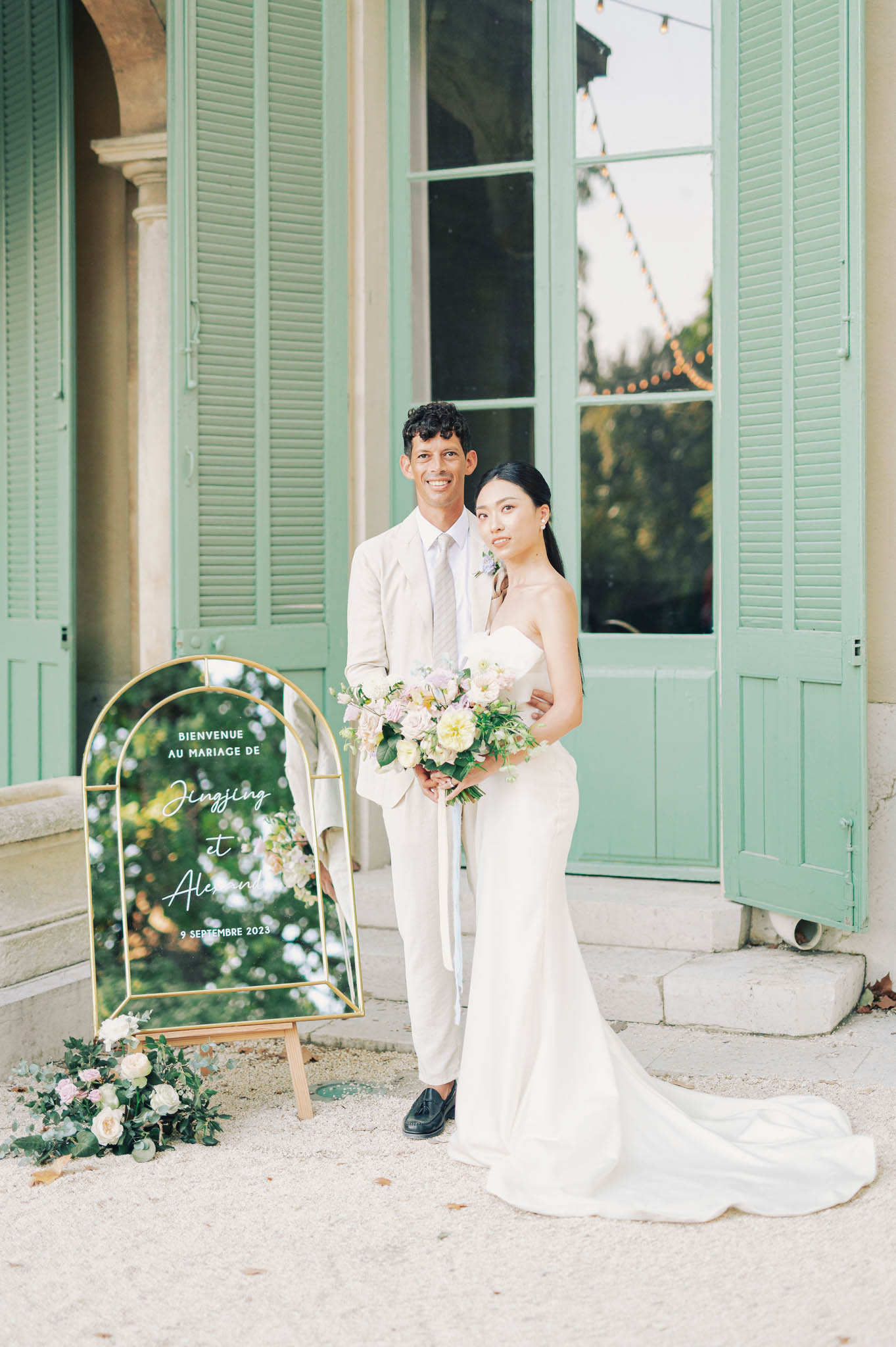 Bride and groom posing in front of chateau with green shutters beside gold mirror welcome sign
