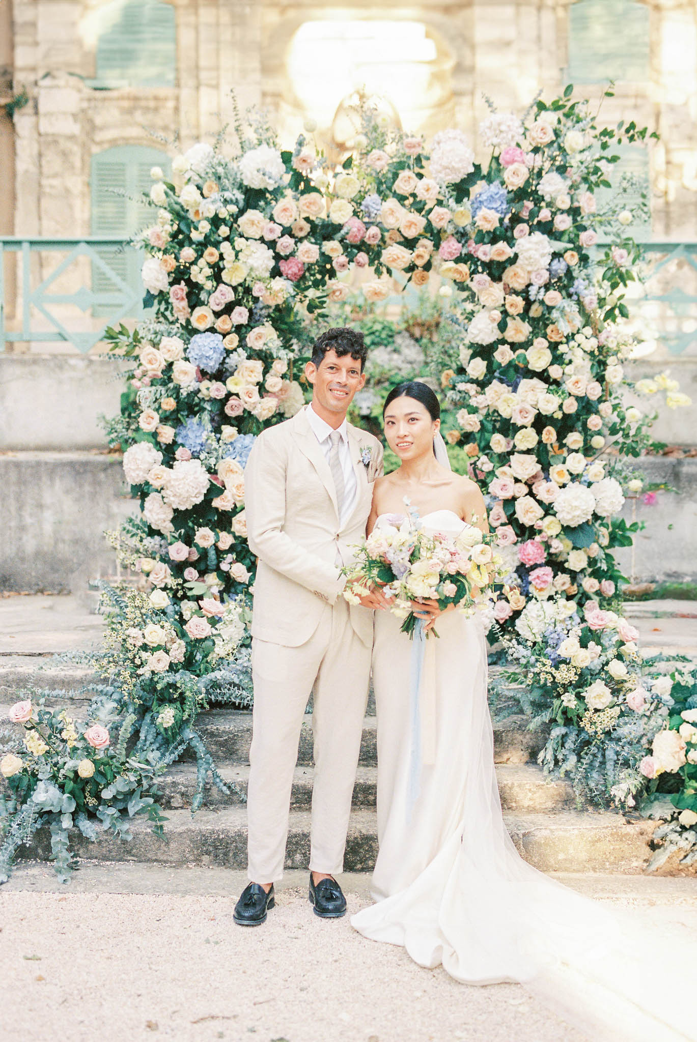 A couple portrait taken outdoors in front of a large circular floral arch installed against the stone façade of what appears to be a French château, with teal-painted shutters visible in the background. The groom wears a light sand/beige suit with a silver-grey tie and black tassel loafers, while the bride wears a strapless ivory fitted gown with a long train and a sheer veil; she holds a loose bouquet of cream, blush, and pale yellow roses with blue accents. The circular arch is densely arranged with cream garden roses, blush and peach roses, blue and white hydrangeas, pink blooms, and lush greenery, with additional ground-level floral arrangements of eucalyptus, blush roses, and wildflower-style stems flanking the base. The overall floral palette is soft pastel — cream, blush, peach, periwinkle blue, and pale yellow — styled in a romantic classic French manner, captured as a mid-length couple portrait in natural warm light.