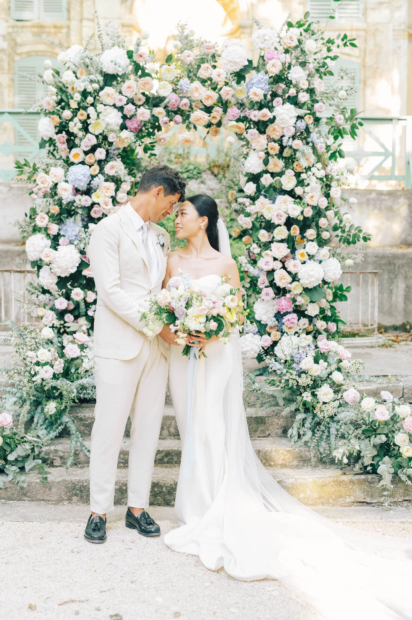 Bride and groom standing forehead to forehead beneath a circular floral arch of hydrangeas and roses on chateau stone steps