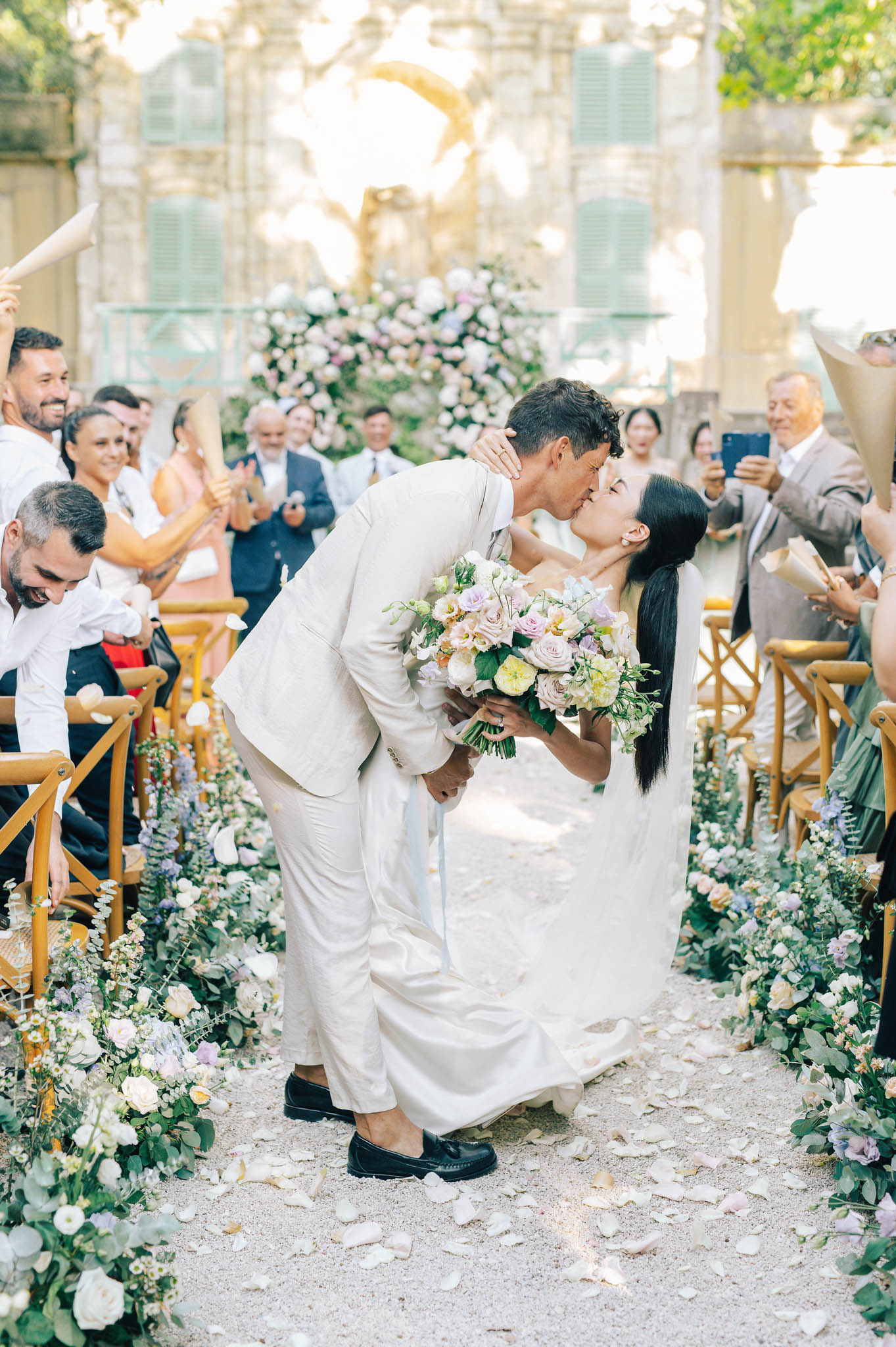 Couple shares dip kiss mid-aisle after outdoor ceremony in stone courtyard with floral arch behind