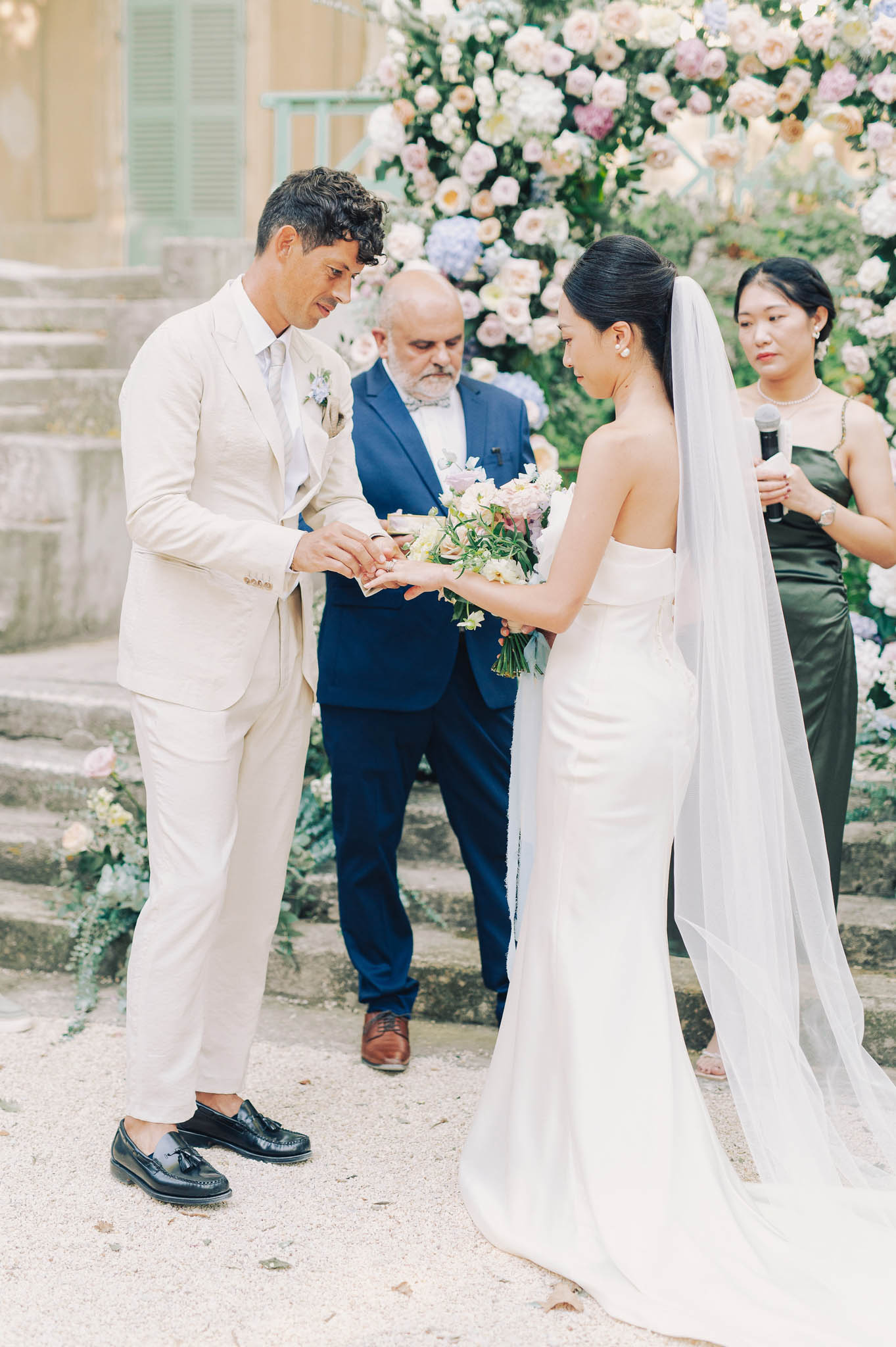 Ring exchange during outdoor ceremony with floral arch of blush roses and lavender hydrangeas at chateau steps