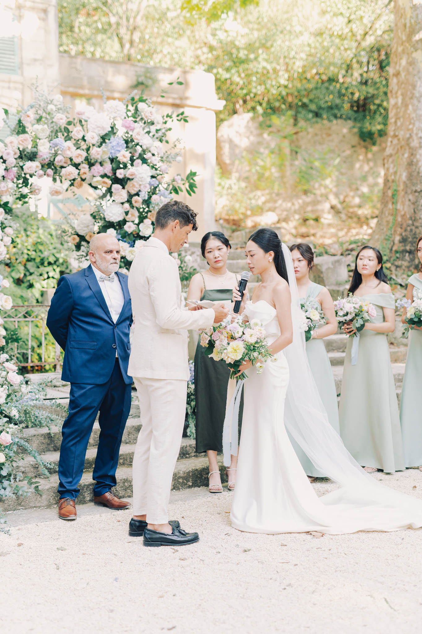 An outdoor wedding ceremony vow exchange taking place in the grounds of what appears to be a French stone estate or château, with classical stone steps and architectural columns visible in the background. The bride, wearing a white fitted strapless gown with a long flowing veil and cathedral train, holds a microphone and reads her vows while the groom, dressed in a cream/ivory suit with black loafers, holds her hand and looks down; she carries a loose bridal bouquet of blush, peach, pale yellow, and lilac blooms. Behind the couple stands a large floral installation composed of blush and cream roses, lavender hydrangeas, and lush greenery, while three bridesmaids in sage green off-the-shoulder midi dresses hold matching smaller bouquets, and a woman in an olive green dress stands closest to the couple; an older man in a navy blue suit with a bow tie observes from the left. The composition is a medium full shot with a softly blurred background, giving the image a light, airy feel consistent with a classic French garden wedding aesthetic.