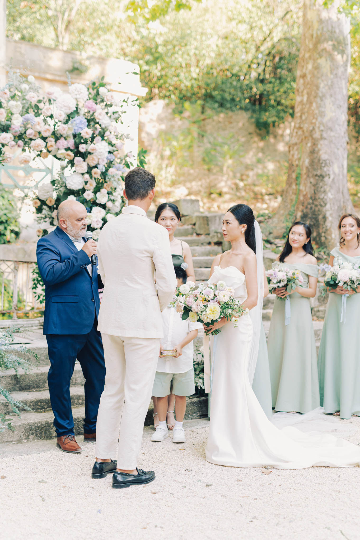 An outdoor wedding ceremony is taking place in a garden setting with stone steps and mature trees in the background. The bride, wearing a strapless fitted white satin gown with a long train and a veil, holds a loose bouquet of soft yellow, blush, and lilac dahlias, ranunculus, and greenery, while facing the groom, who wears a cream linen suit with black loafers. An officiant in a navy blue suit holds a microphone and speaks to the couple, with a small child standing between them holding what appears to be a ring box. Two bridesmaids in sage green off-the-shoulder floor-length dresses hold matching bouquets of blush and lilac blooms, and a third attendant in a black dress stands nearby. A large floral installation featuring blush, ivory, and lavender roses, hydrangeas, and lush greenery is visible on the left beside a white column. The image is a medium-wide portrait-oriented shot with soft, bright natural light.