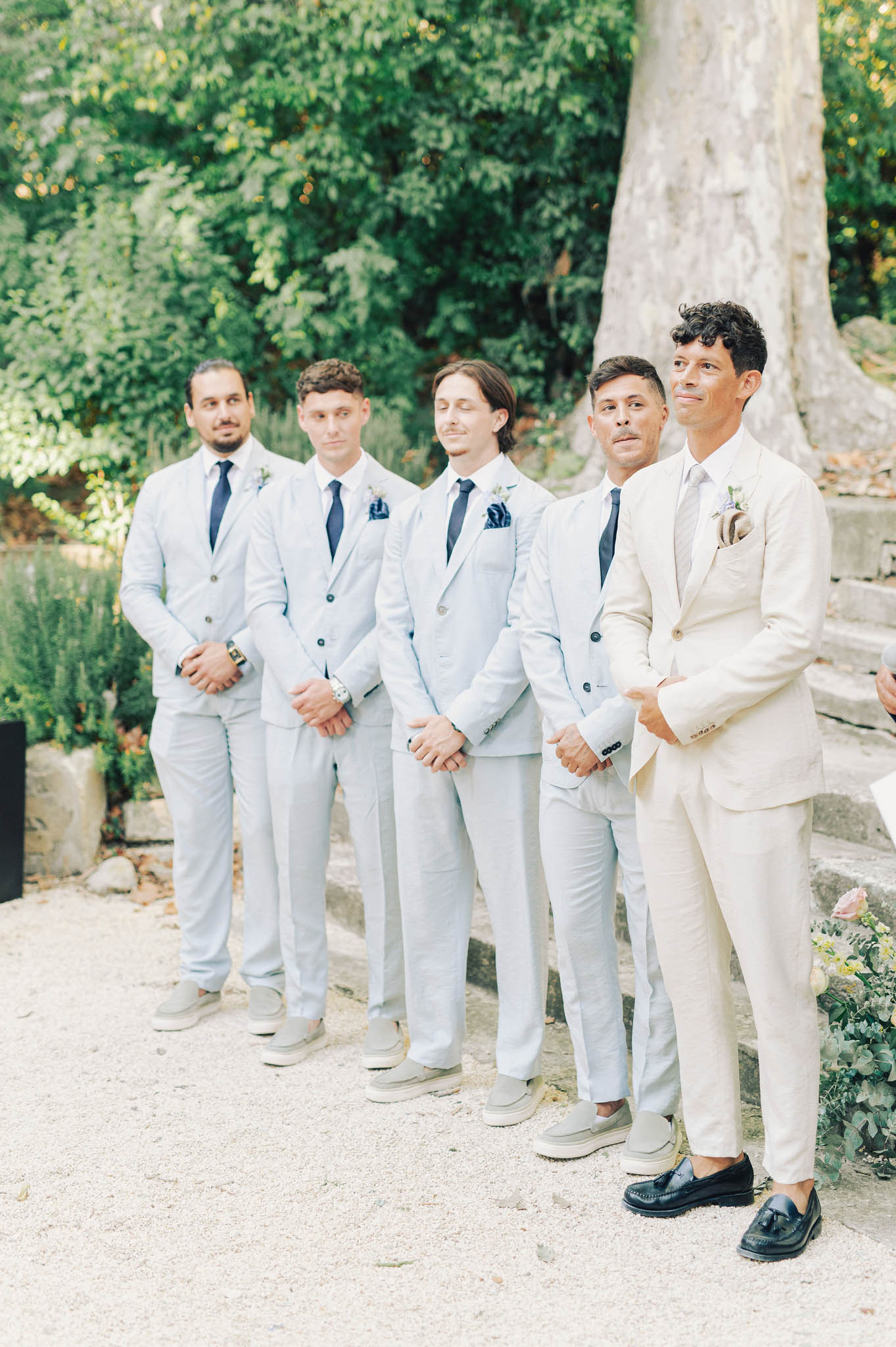 Groom in cream linen suit and four groomsmen in light blue suits with navy ties at outdoor ceremony