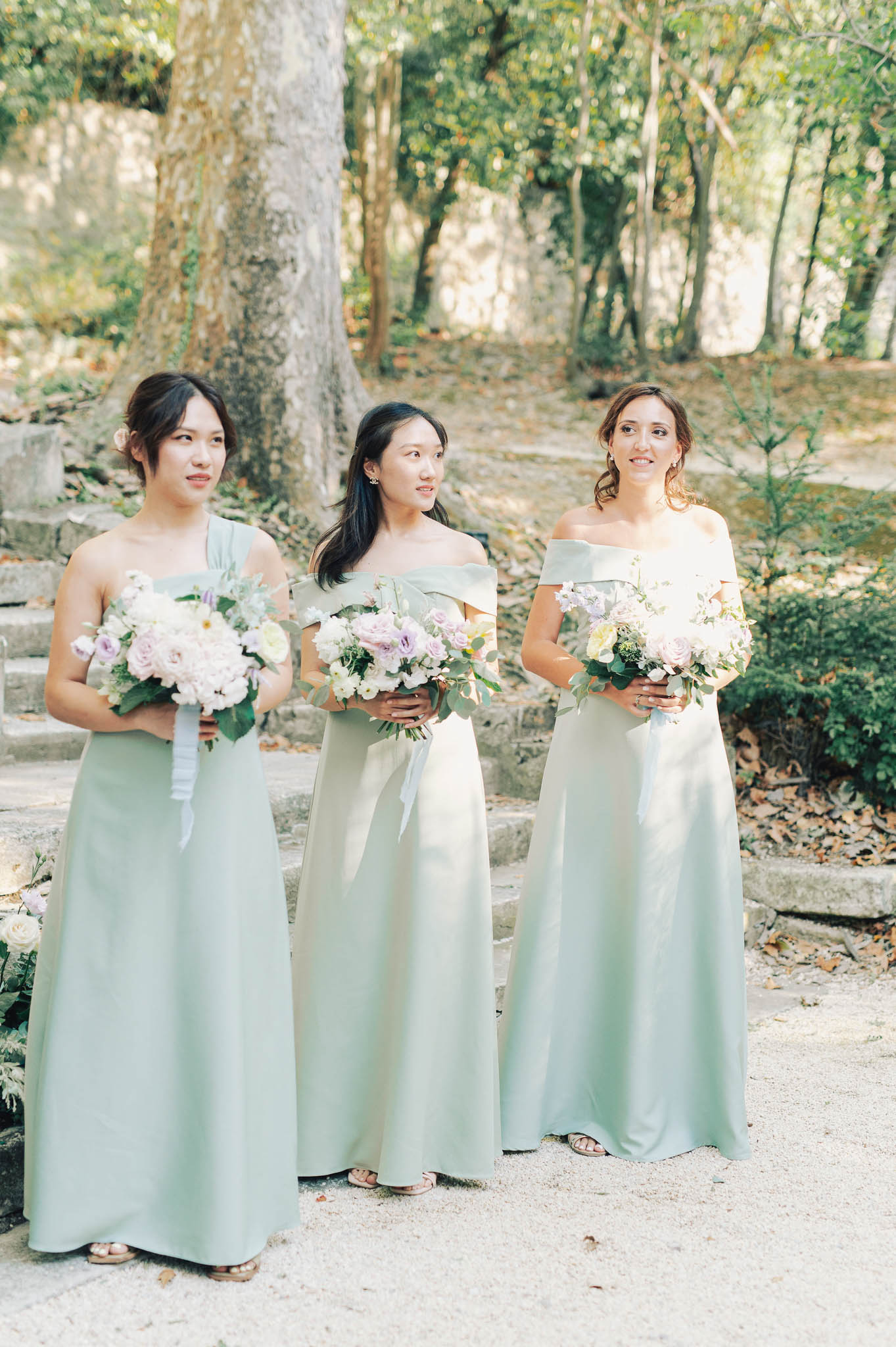 Three bridesmaids stand together outdoors on a gravel path, each holding a bouquet and looking in slightly different directions, suggesting they are positioned during a ceremony procession or bridal party portrait session. The setting features stone steps and a wooded backdrop. All three wear floor-length sage green satin gowns in varying neckline styles — one one-shoulder, two off-the-shoulder — creating a coordinated but slightly mixed look. Each bridesmaid holds a lush bouquet of soft lavender and blush roses, white blooms, and eucalyptus greenery, with white ribbon streamers visible on one bouquet. The image is a mid-shot portrait with soft, natural dappled light and a classic, clean aesthetic.