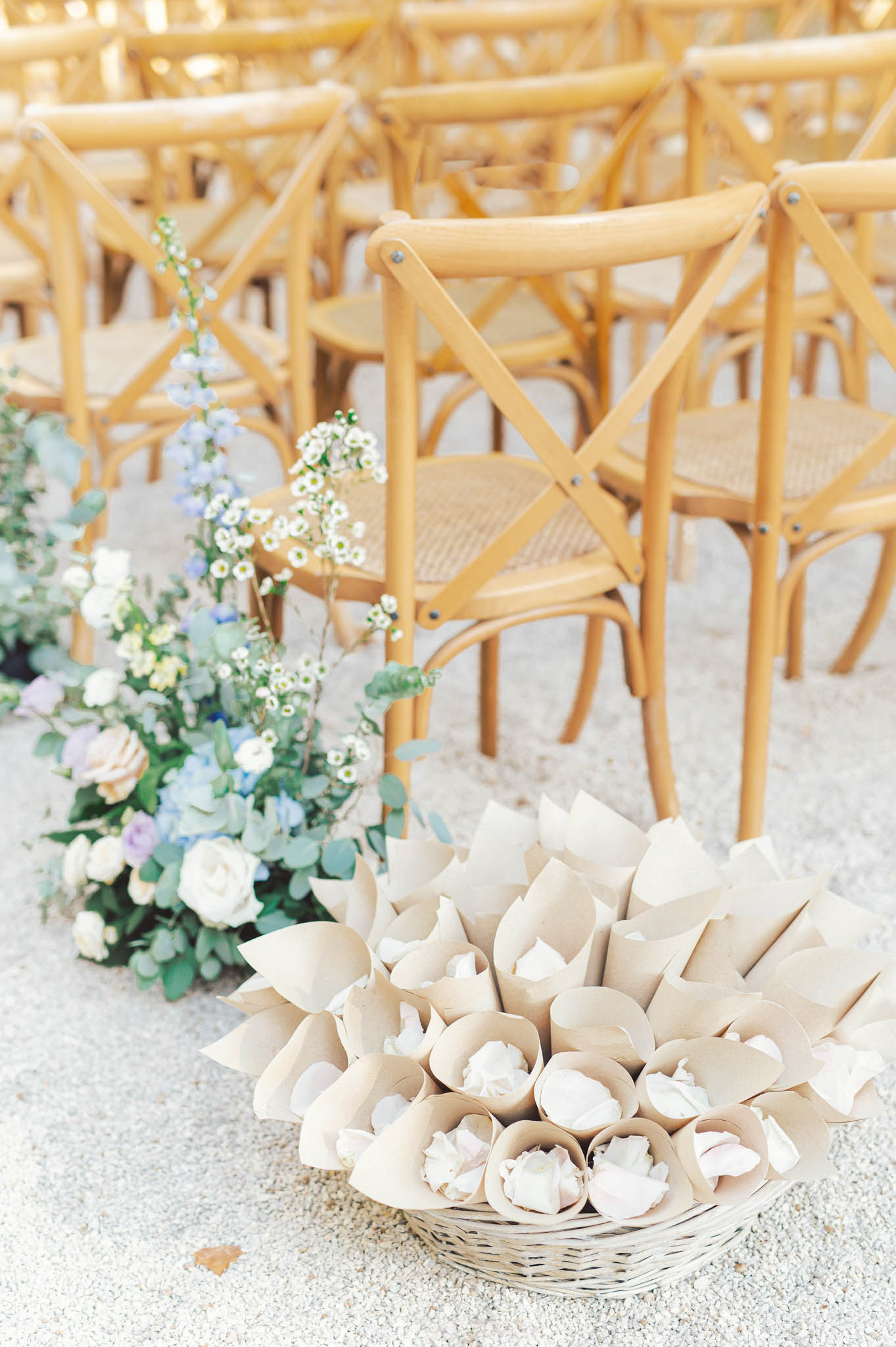 Wicker basket of kraft confetti cones beside blush rose, blue hydrangea, and delphinium aisle arrangement