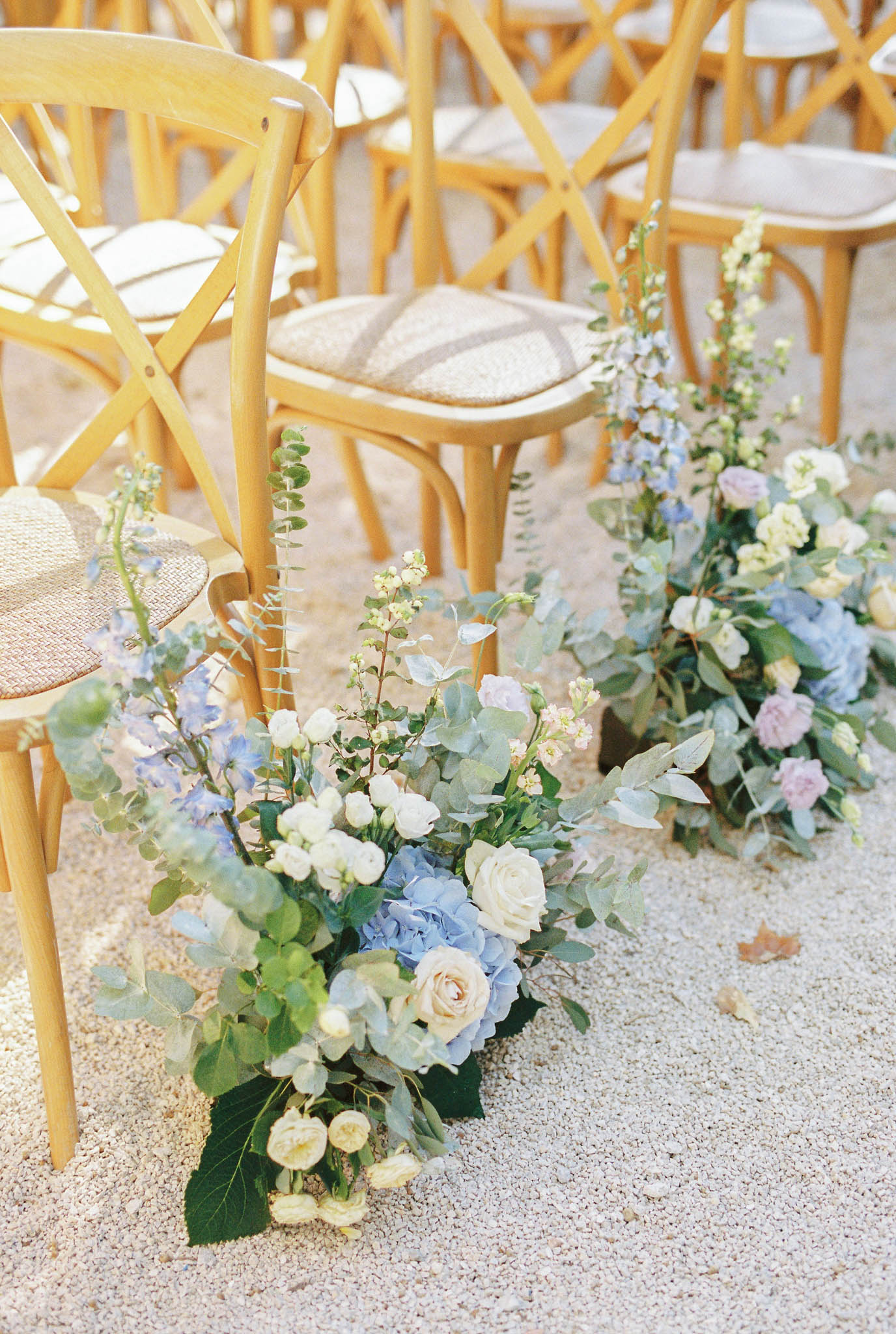 Close-up detail shot of outdoor wedding ceremony aisle décor placed on a gravel surface. Two floral arrangements are positioned along the aisle beside natural wood cross-back chairs with woven rattan seats. The foreground arrangement features pale blue hydrangeas, cream and blush garden roses, white stock flowers, blue delphiniums, and eucalyptus foliage in a loose, garden-style composition. The background arrangement echoes the same palette with soft lavender blooms and additional hydrangeas. The overall floral color palette is powder blue, cream, blush, and soft lavender with abundant green foliage. No people are present in the image.