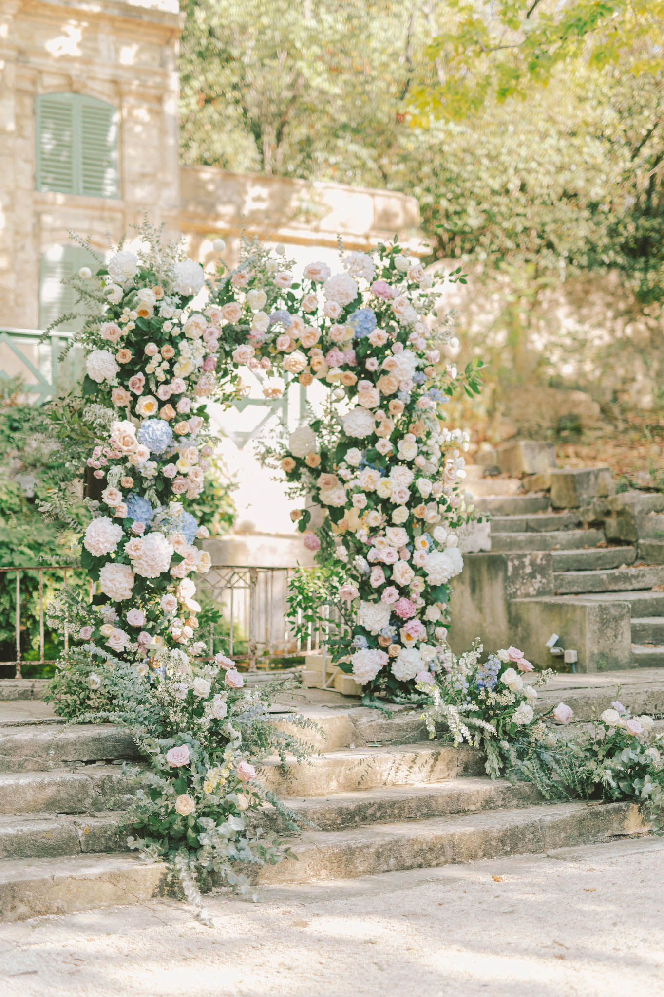 Circular floral arch with blush roses peach peonies and blue hydrangeas atop stone steps at French estate