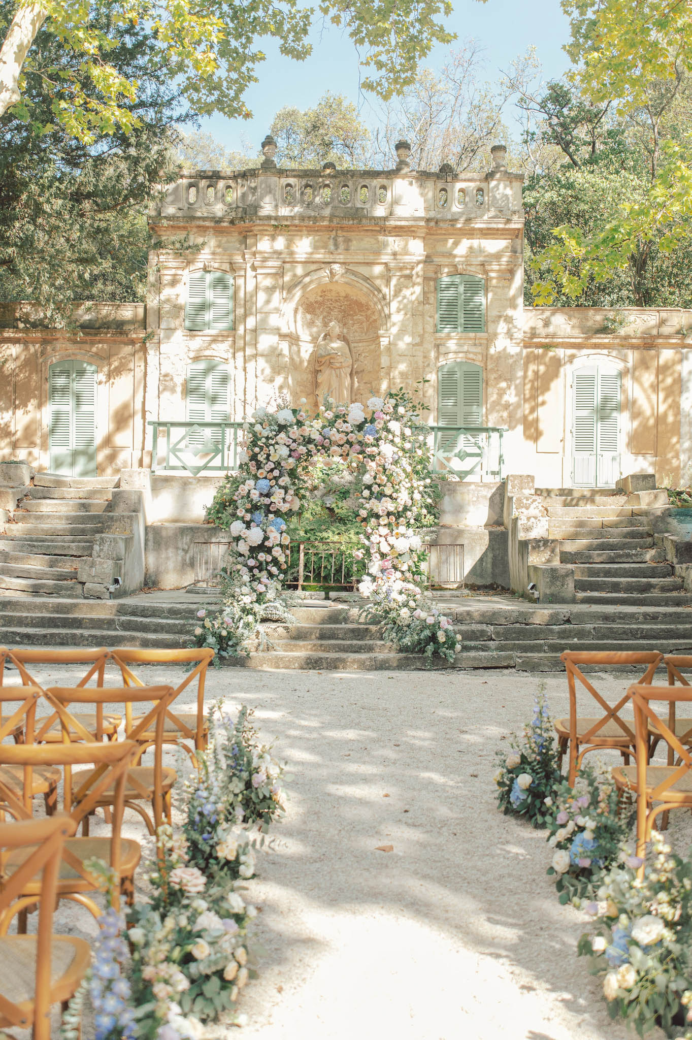 An outdoor ceremony setup photographed from the aisle looking toward the altar, with no people present. The setting is the courtyard of a historic French stone building featuring sage green shutters, a central stone niche with a classical sculpture, and symmetrical stone staircases on either side. A large floral arch frames the altar area, densely arranged with blush pink roses, white hydrangeas, blue hydrangeas, peach garden roses, and trailing greenery. Smaller floral clusters in the same palette — blush, white, soft blue, and peach — line both sides of the gravel aisle at the base of each natural wood cross-back chair. The ceremony chairs are arranged in two sections on either side of the aisle. The overall decor palette is soft and romantic, combining blush, dusty blue, and ivory florals against the warm honey-toned stone architecture. Wide shot taken from ground level down the aisle. Potential venue feature image.