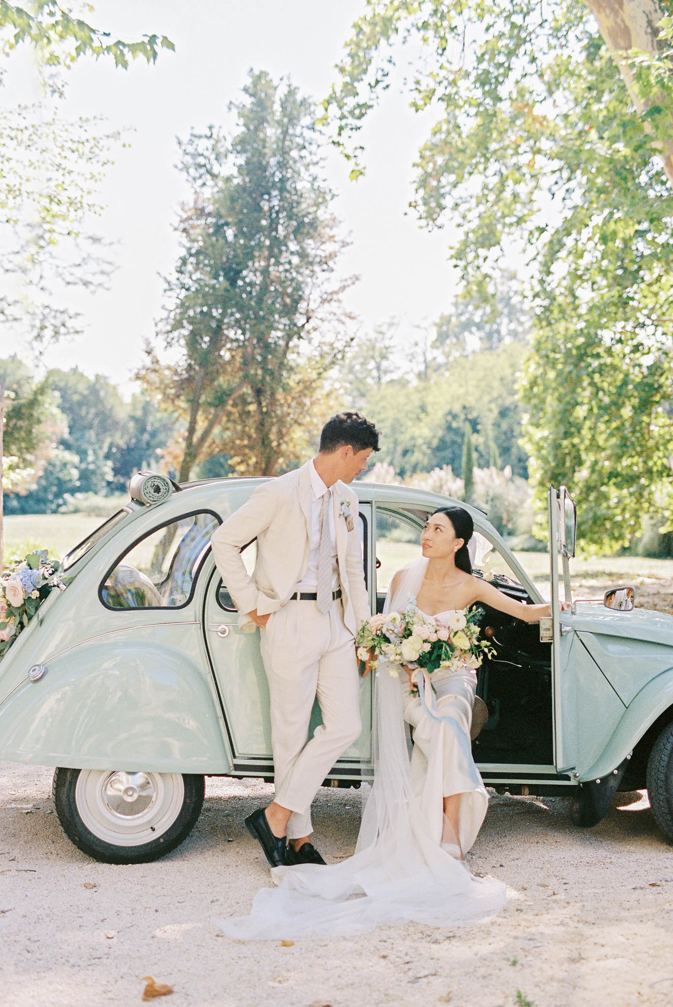 A couple poses outdoors in front of a vintage mint-green Citroën 2CV decorated with small floral clusters including pale blue hydrangeas and blush roses on the front fender. The groom stands leaning against the car wearing a cream linen suit with a matching tie and a small boutonniere, paired with black dress shoes. The bride is seated in the open passenger doorway wearing a strapless ivory gown with a long flowing veil that pools on the ground; she holds a loose, garden-style bouquet featuring cream, blush, and yellow blooms with greenery and trailing ribbons. The two are looking at each other in a gravel courtyard setting surrounded by mature trees. The image is a full-length portrait shot taken in natural daylight with a soft, film-like quality and a warm, airy tone.