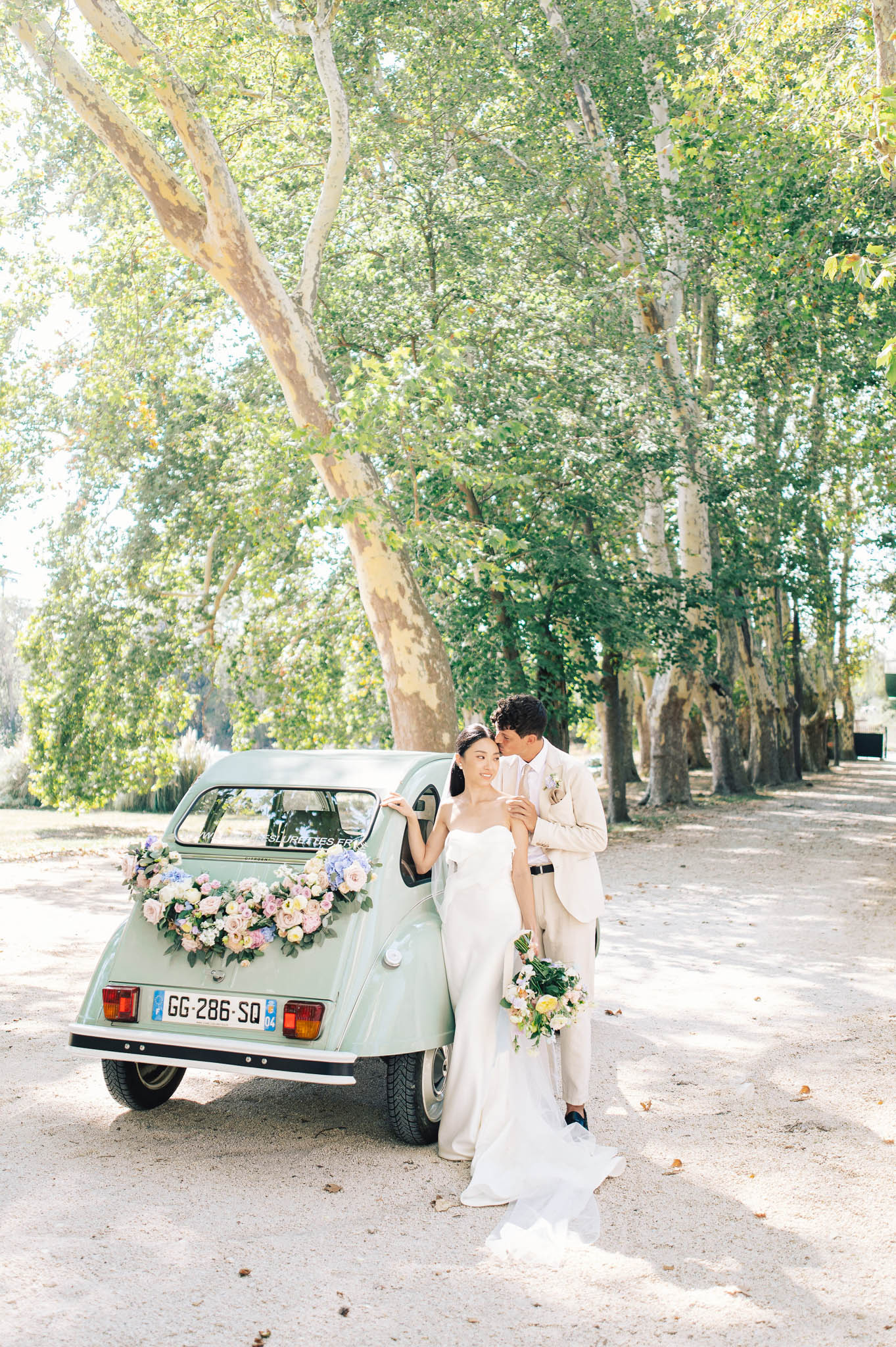 Groom kissing brides cheek beside mint-green 2CV with blush and lavender rose garland on tree-lined gravel drive