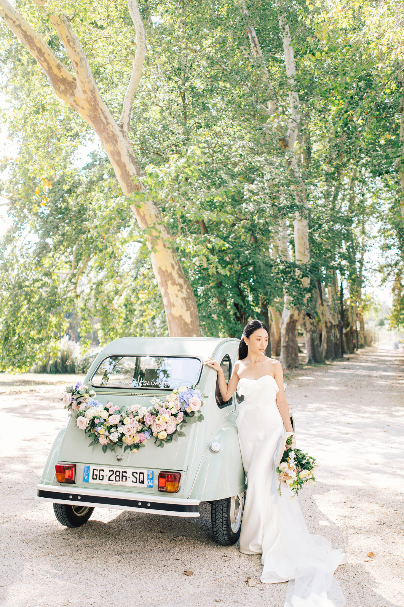 A bride poses leaning against the rear of a vintage mint-green Citroën 2CV on a tree-lined gravel driveway, captured in a full-length portrait shot. The car's boot is decorated with a floral arrangement of blush pink, lavender, and cream roses with eucalyptus foliage. The bride wears a strapless ivory fitted gown with a short train and holds a loose bouquet of peach, cream, and blush blooms with greenery. The setting is an outdoor avenue of large plane trees creating a natural canopy, likely on the grounds of a French estate or château.