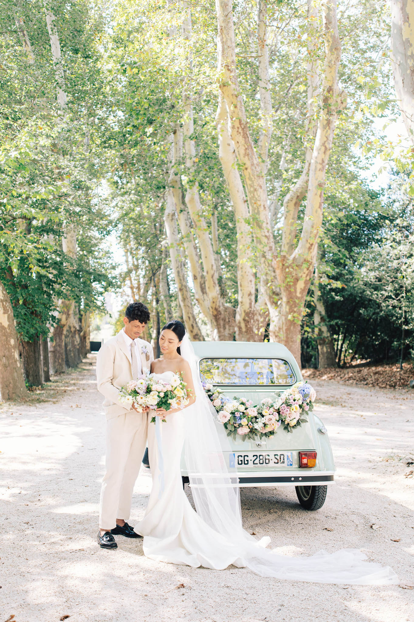 Bride and groom standing by vintage mint-green Citroen 2CV decorated with pink rose garland on tree-lined allee