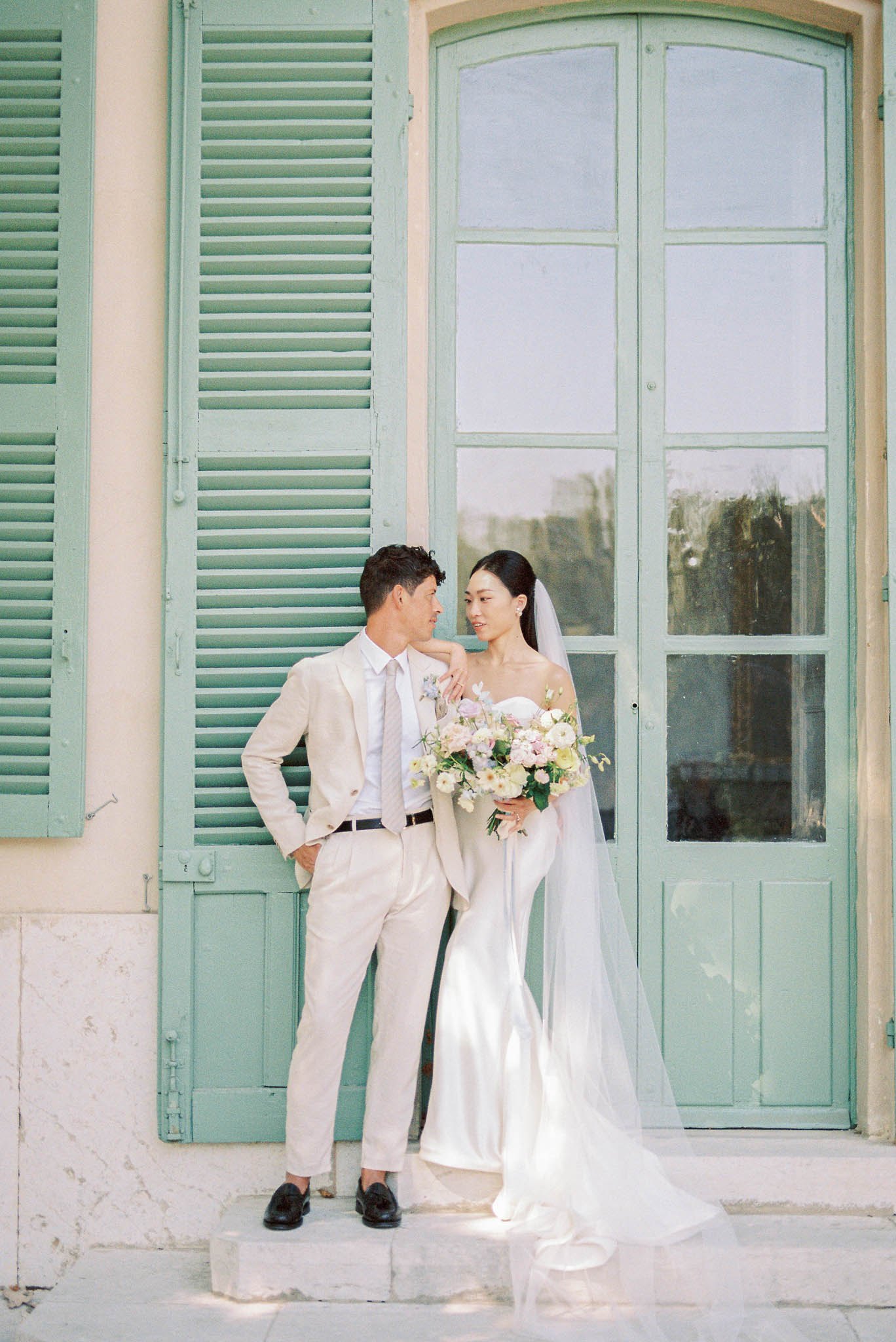 A couple portrait taken outdoors against the facade of a French-style building featuring sage green louvred shutters and a large arched sage green door with glass panels. The groom wears a light sand-colored suit with a white shirt, a pale striped tie, a small floral boutonnière in soft lavender and white, and black loafers; he leans casually against the shutters with one hand in his pocket. The bride wears a fitted ivory strapless satin gown with a long flowing train and a floor-length veil; she rests one hand on the groom's shoulder and holds a loose, garden-style bouquet of cream ranunculus, blush roses, soft yellow blooms, and delicate wildflowers with trailing greenery. The two are looking at each other in a relaxed, close pose. The overall styling is modern and clean with a soft, warm color palette. Medium portrait shot with the building architecture filling the background. Potential venue feature image.